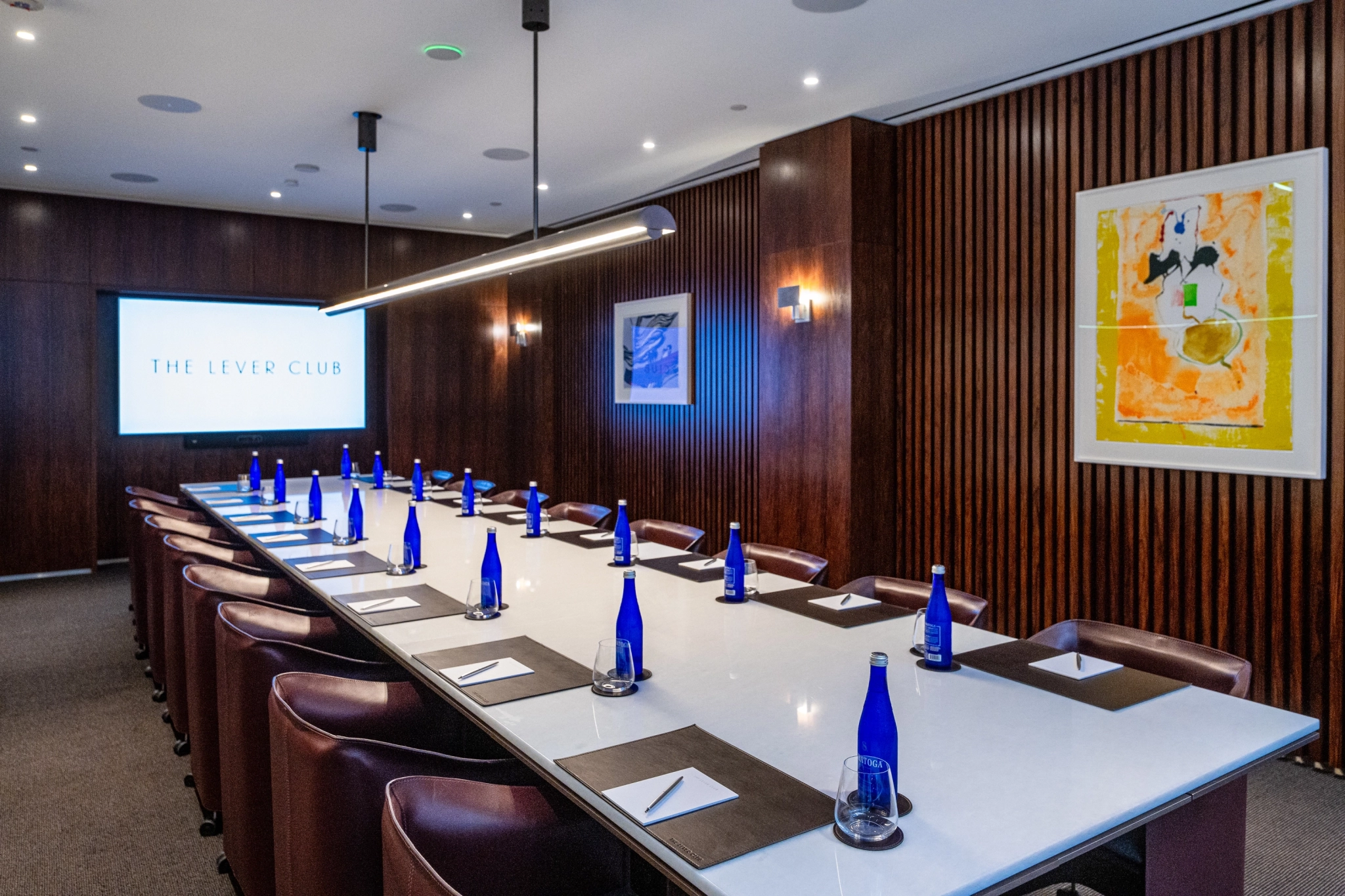 Una moderna sala de reuniones en Nueva York con una larga mesa blanca, sillas de cuero, botellas de agua azules, cuadernos y una gran pantalla en la que se proyecta "The Lever Club".
