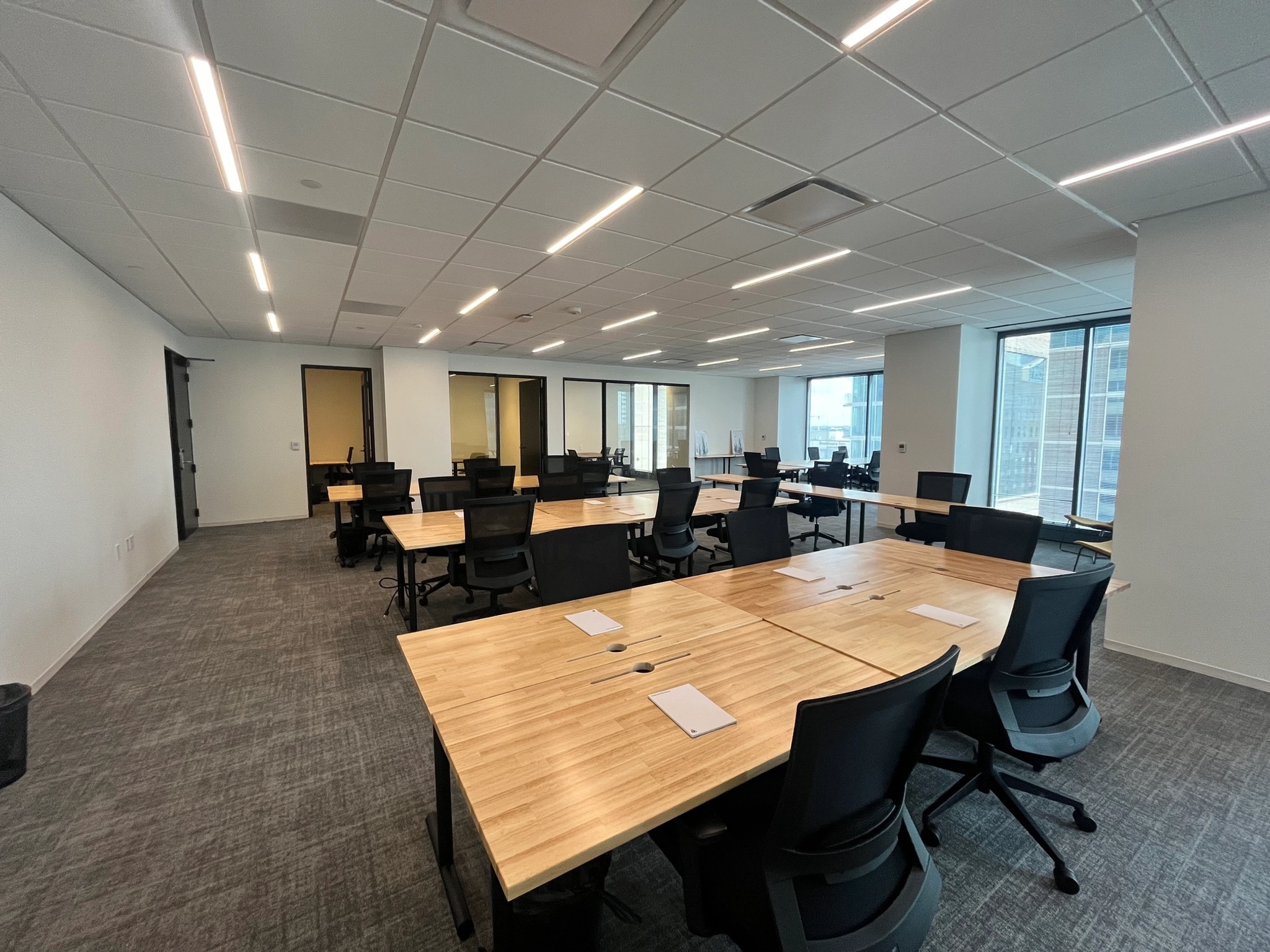 A modern open-space coworking office in Austin features several wooden desks and black chairs, arranged in rows. Large windows on the far side allow natural light to fill this vibrant workspace.