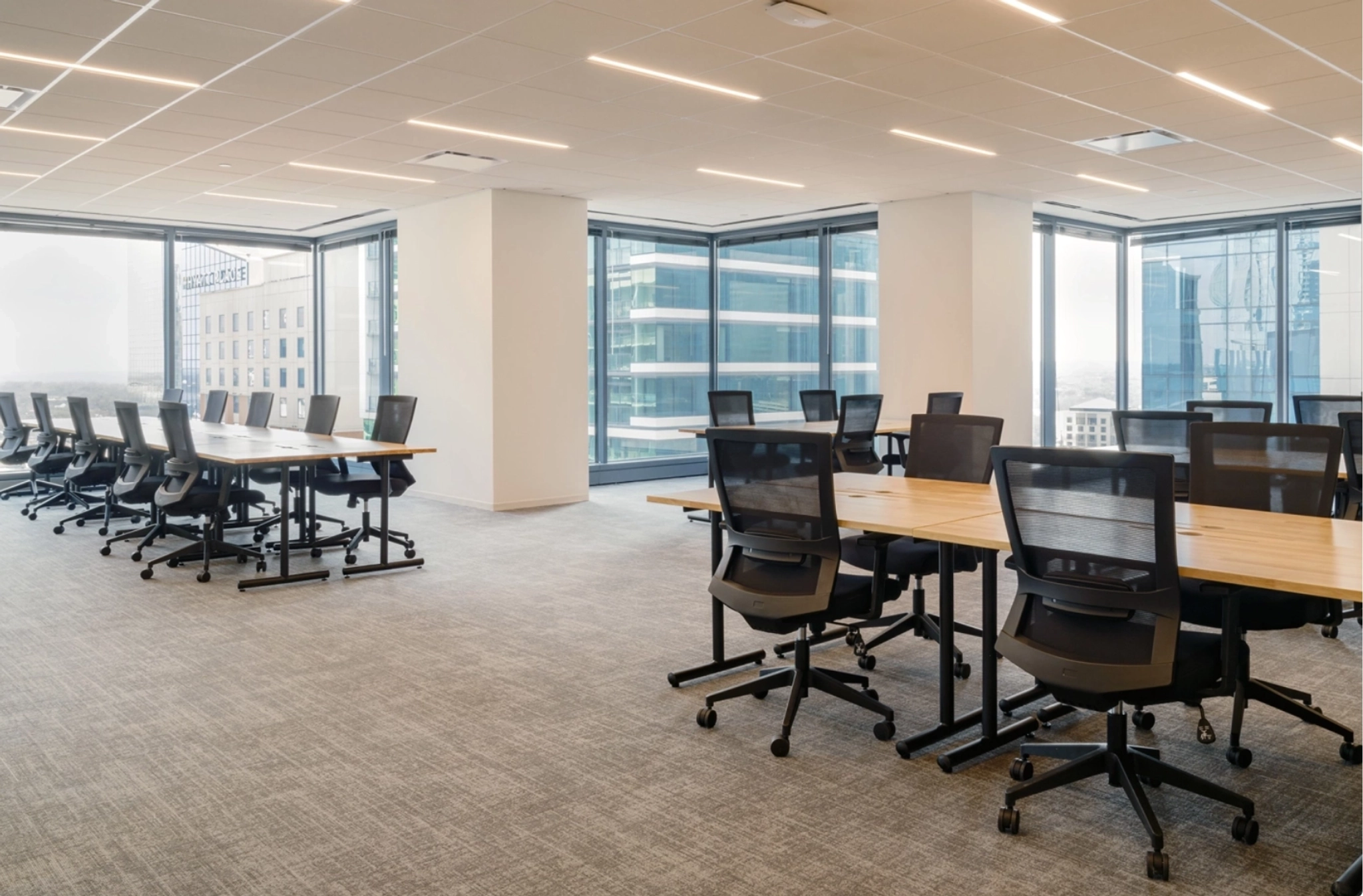 A modern, spacious workspace with multiple desks and chairs, large windows offering a view of adjacent buildings. The floor is carpeted, and the ceiling features recessed lighting, creating an inviting office environment.