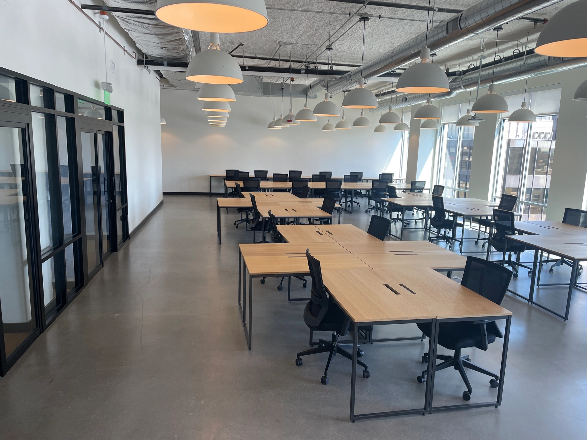 Located in the heart of Seattle, this modern coworking office space boasts large wooden desks, black chairs, and overhead pendant lights. With expansive windows along one wall and a glass-walled meeting room on the left side, it offers an ideal environment for collaboration and productivity.