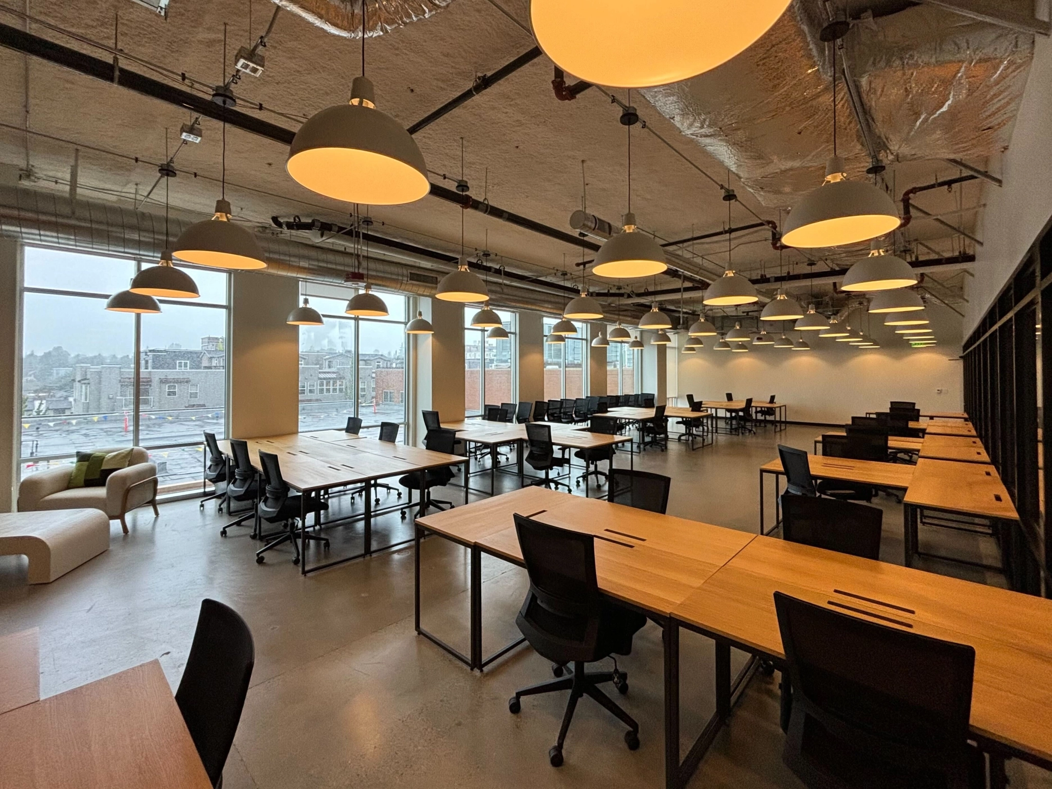 Un bureau de coworking spacieux et moderne à Seattle avec plusieurs bureaux et chaises en bois, un éclairage lumineux au plafond, de grandes fenêtres et un plafond industriel.