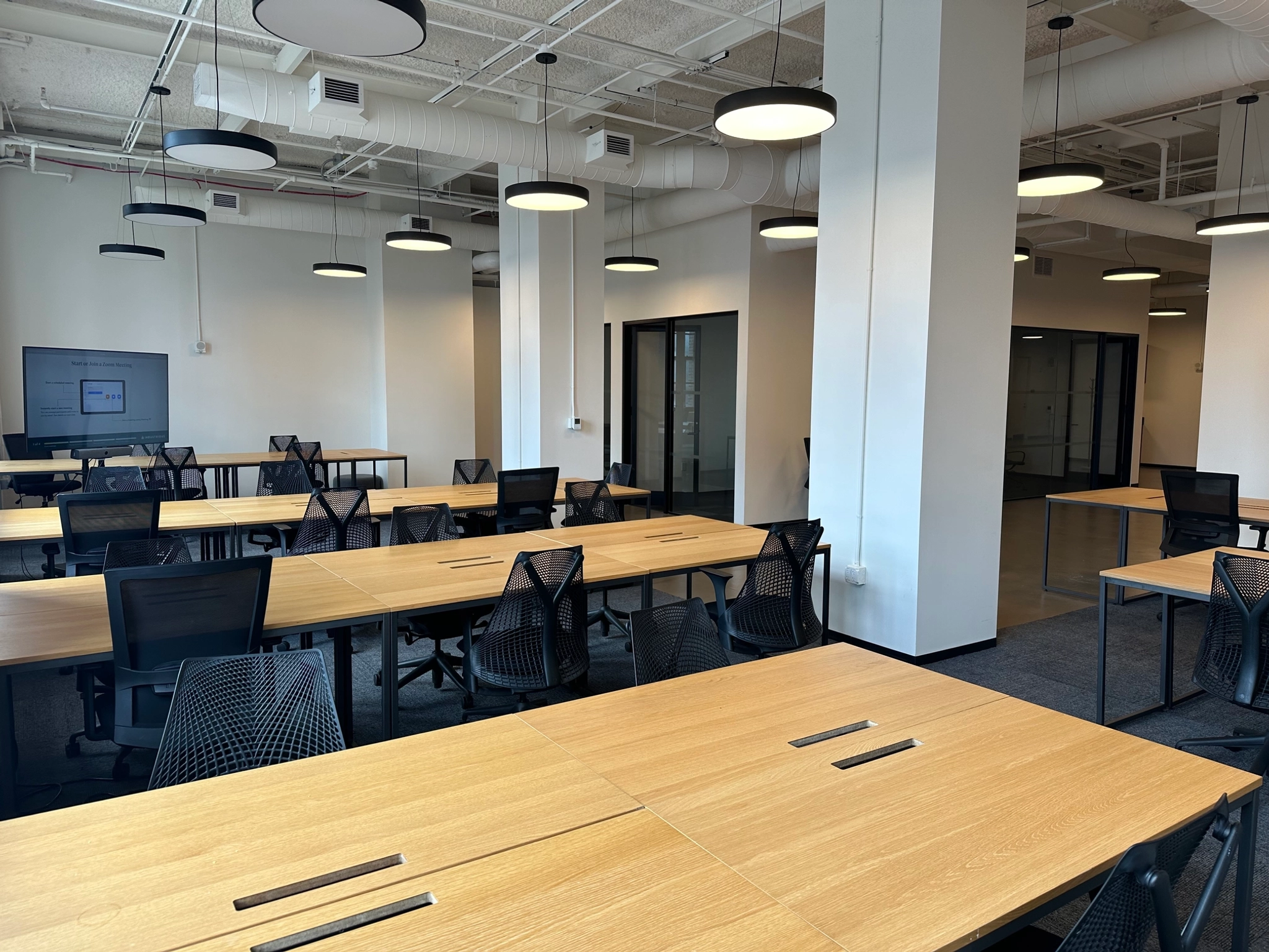 A spacious coworking area with multiple workstations featuring wooden desks and black mesh chairs. The workspace is well-lit with ceiling lights and has a modern industrial design with exposed ducts and white walls.