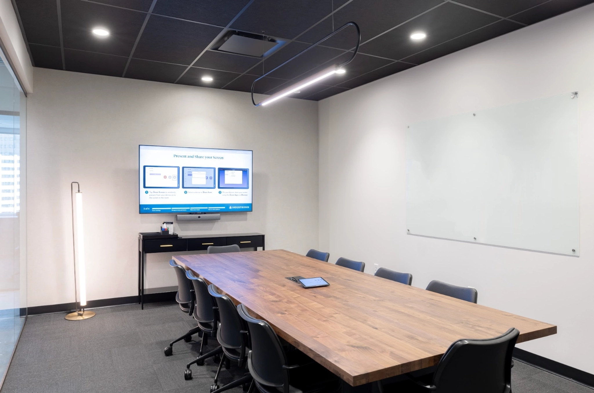 A modern conference room in the coworking space features a wooden table, black chairs, a wall-mounted screen displaying a presentation, a whiteboard, and a tall floor lamp. A tablet is placed on the table, making it an ideal workspace for collaboration.