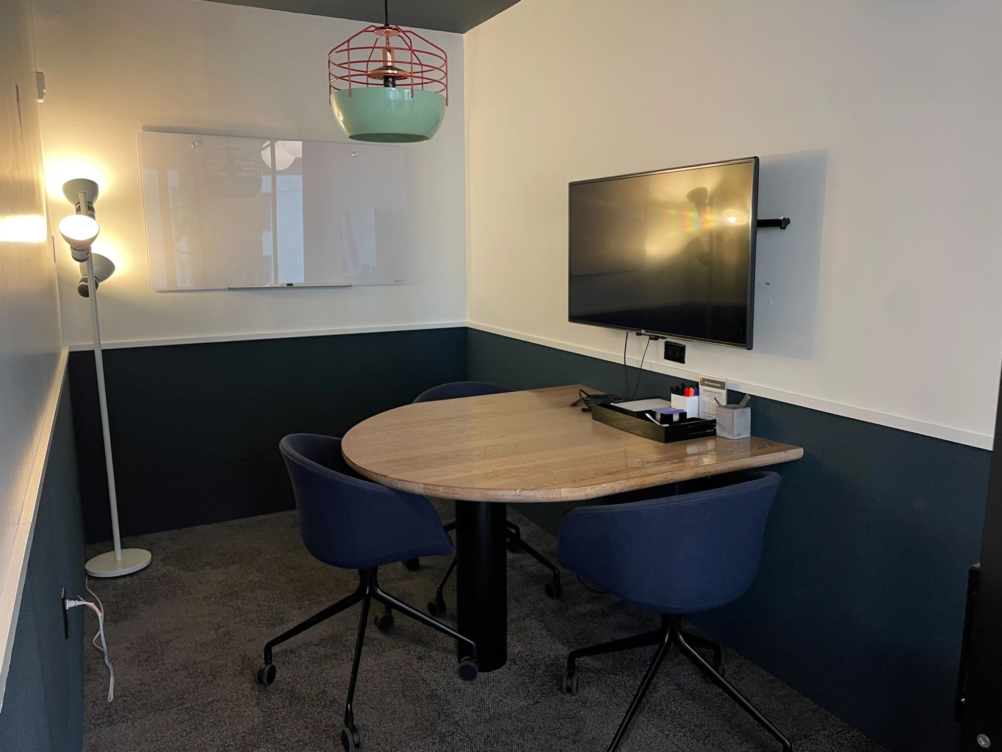 A small coworking meeting room with a round table, four chairs, a mounted TV, a whiteboard, and a floor lamp. The office space features a two-tone wall design, with the lower half being dark and the upper half light-colored.