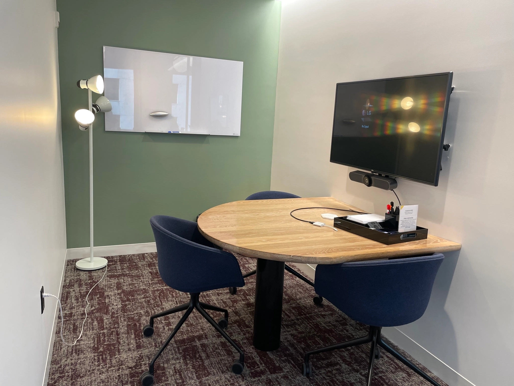 Gezellige vergaderruimte in New York met een ronde houten tafel, twee blauwe stoelen, een tv aan de muur, een whiteboard, een staande lamp en stroomkabels op een tapijt met bruin patroon - perfect voor je volgende werkvergadering.