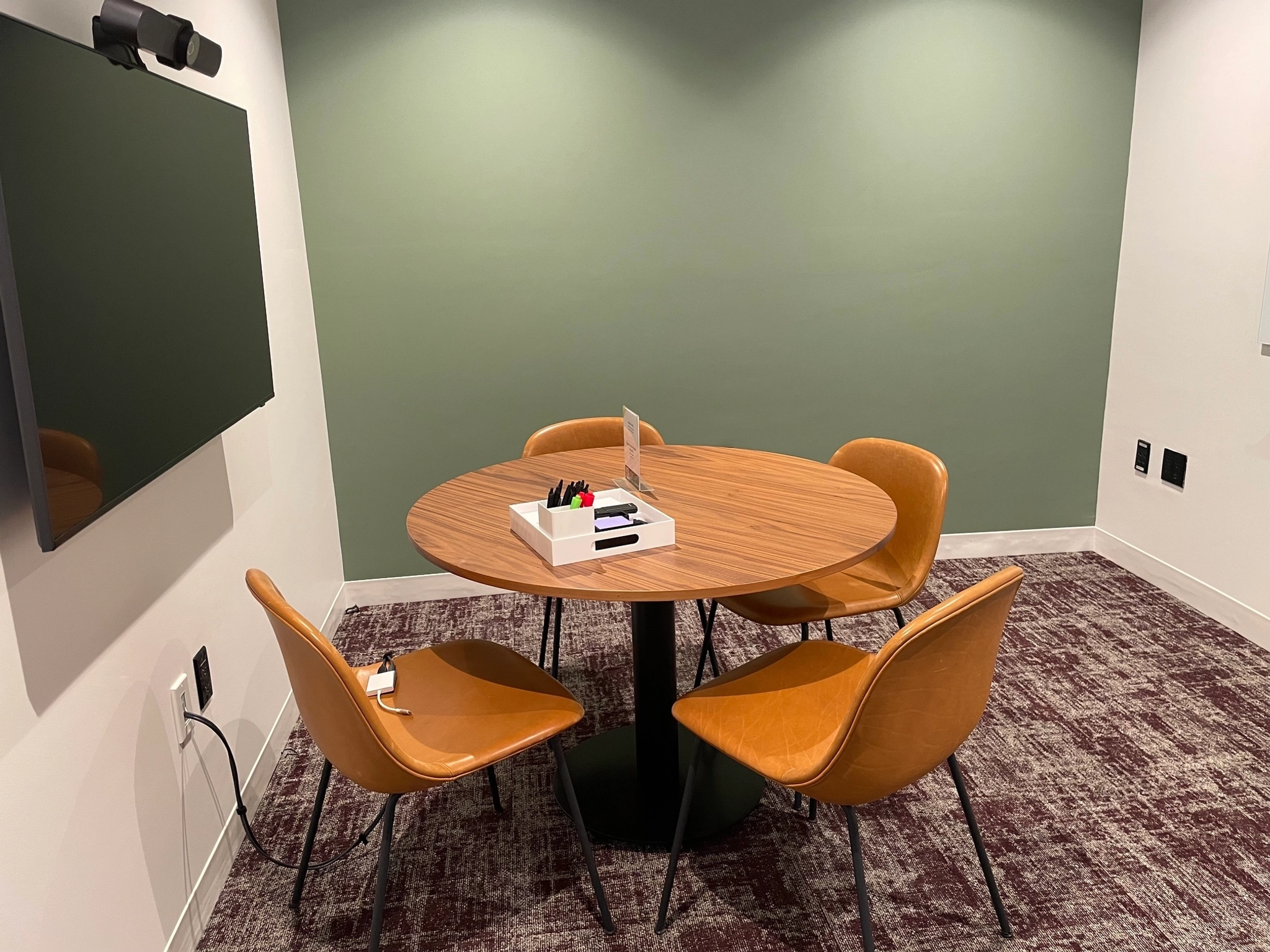 Een kleine coworking vergaderruimte in New York met een ronde houten tafel, vier leren stoelen, een scherm aan de muur en kantoorbenodigdheden netjes opgeborgen in een dienblad op tafel.