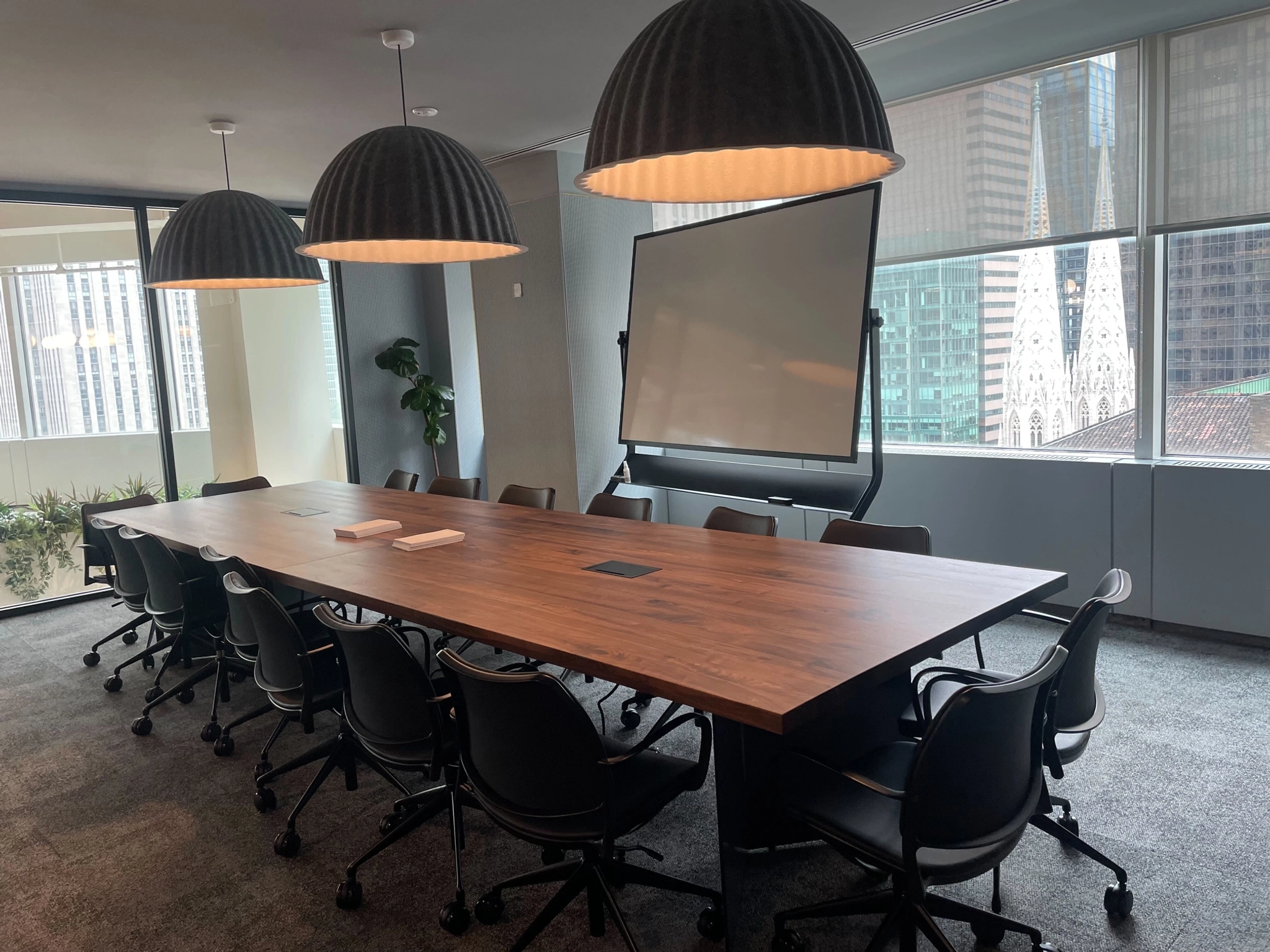 A modern meeting room with a long wooden table, ten black chairs, three large pendant lights, a flip chart, and large windows showcasing New York cityscape views.