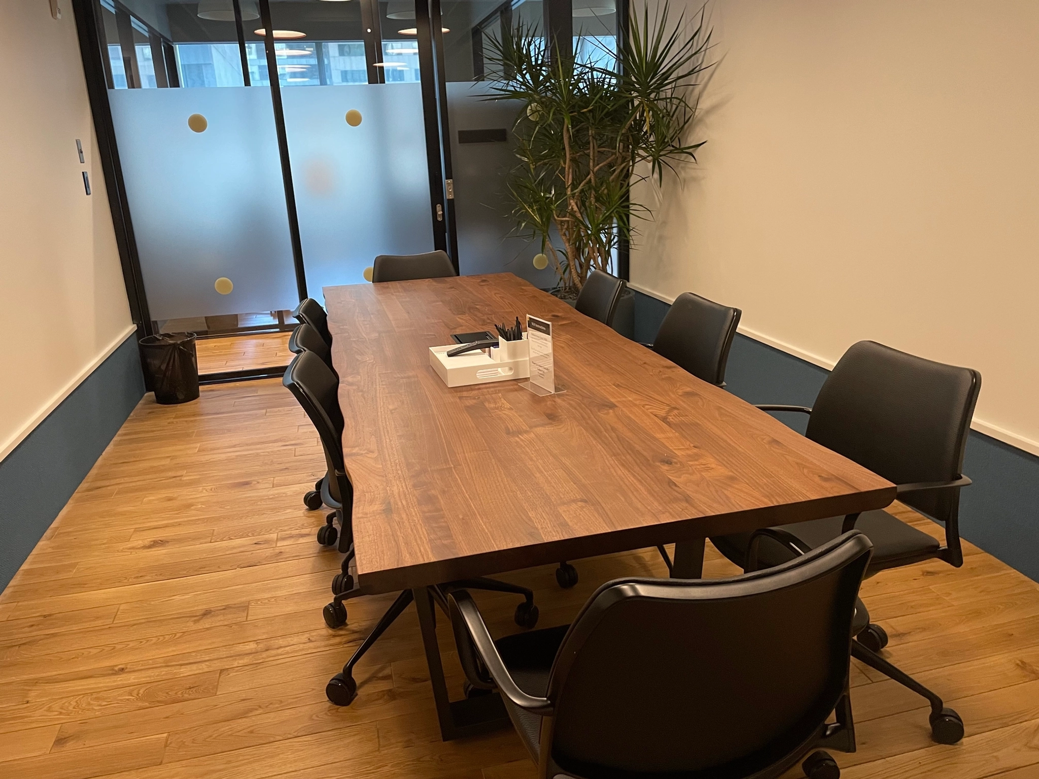 Een stijlvolle vergaderruimte in New York beschikt over een houten tafel, acht zwarte stoelen, een plant in de hoek en een glazen wand met matte panelen, perfect voor dynamische coworking-sessies.