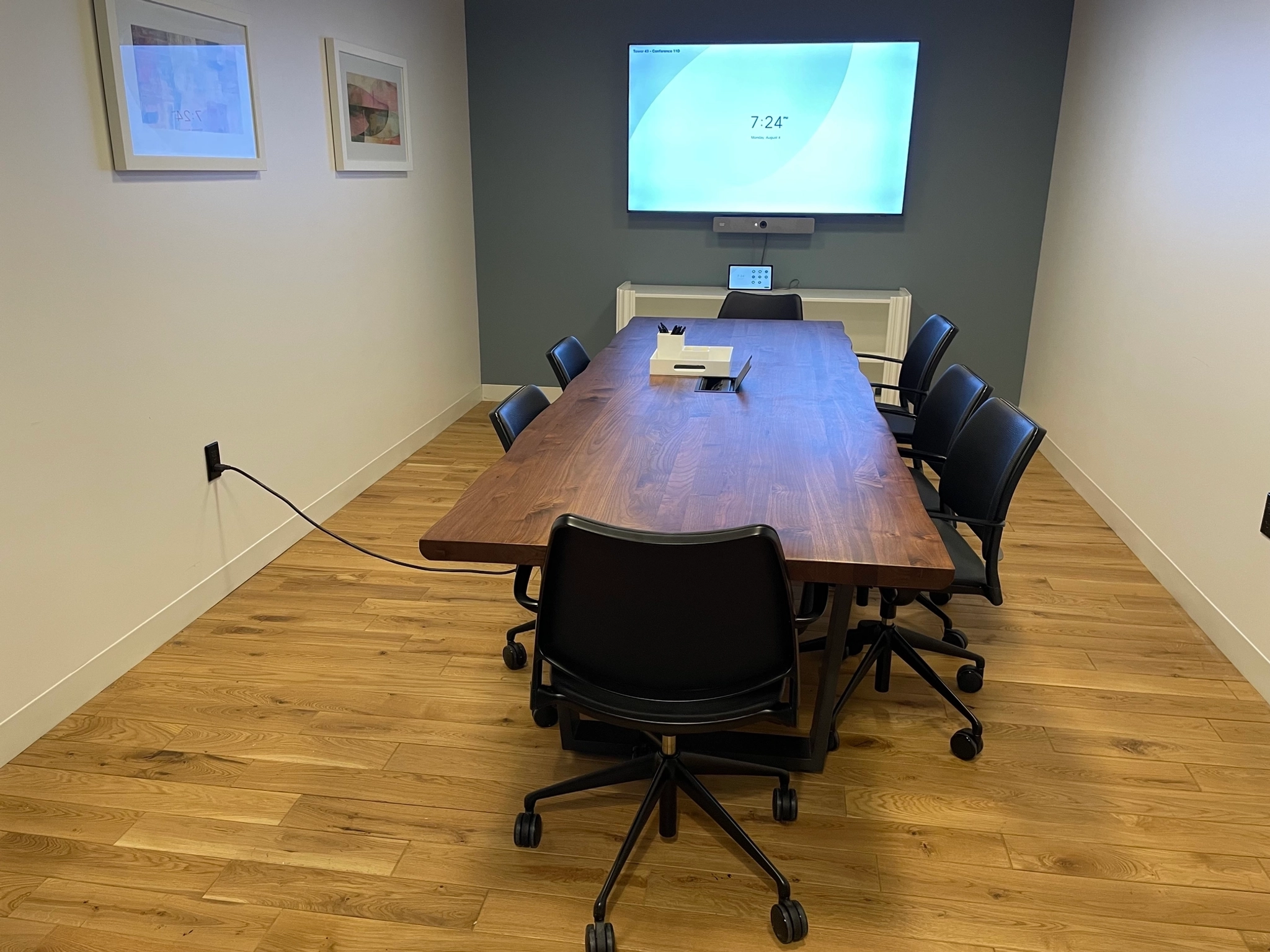 A modern meeting room with a wooden table, six black chairs, a wall-mounted screen showing 72.4°F, two framed pictures, and a device on the table—perfect for any coworking or office environment.