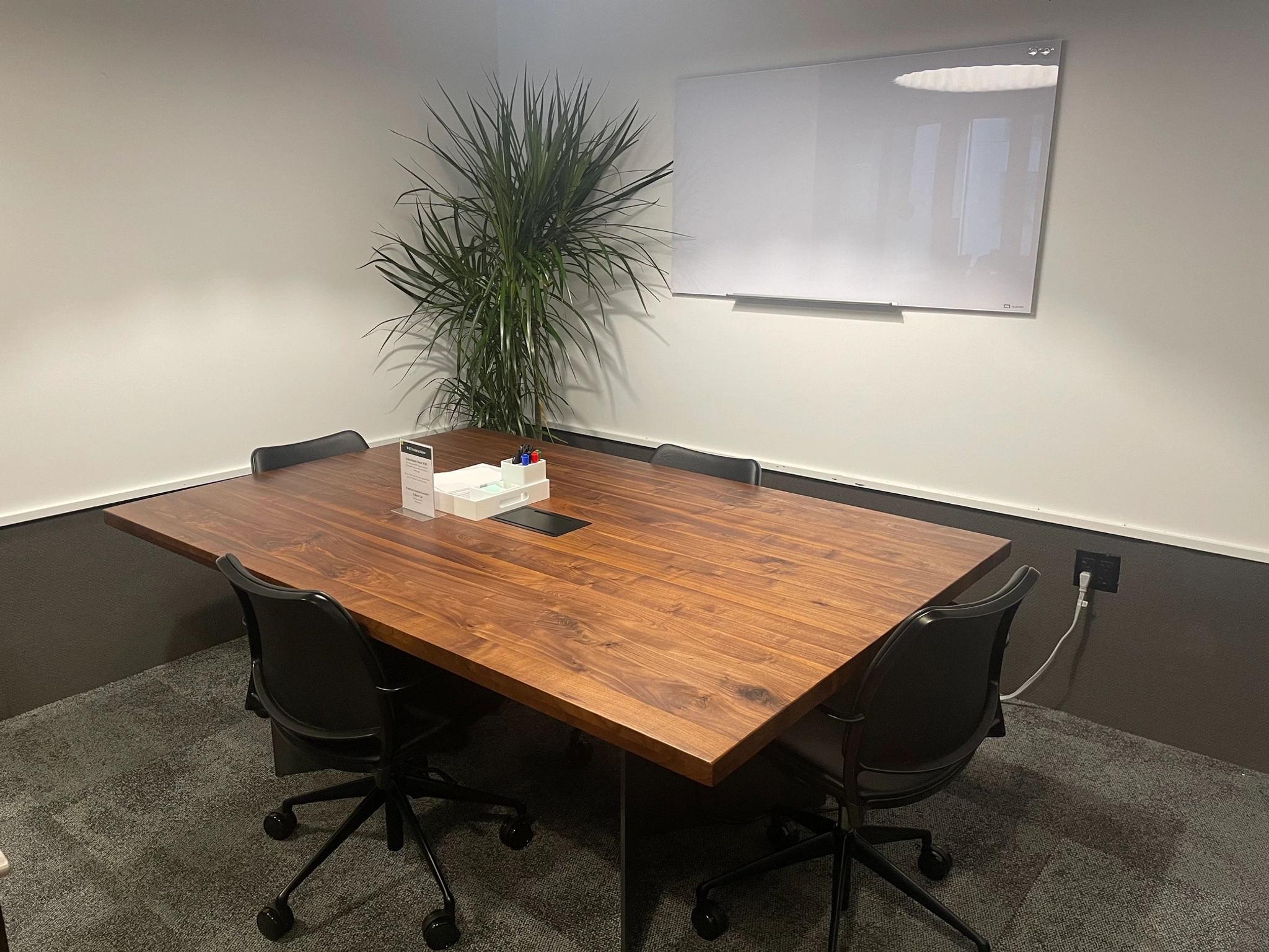 A meeting room with a wooden table, six chairs, a potted plant, a tissue box, markers, and a whiteboard on the wall provides an ideal workspace for collaboration.