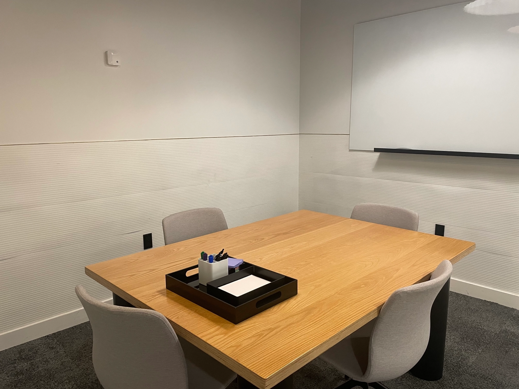 A cozy meeting room with a wooden table, four gray chairs, and a whiteboard on the wall. This coworking space includes a black organizer filled with stationery for your convenience.