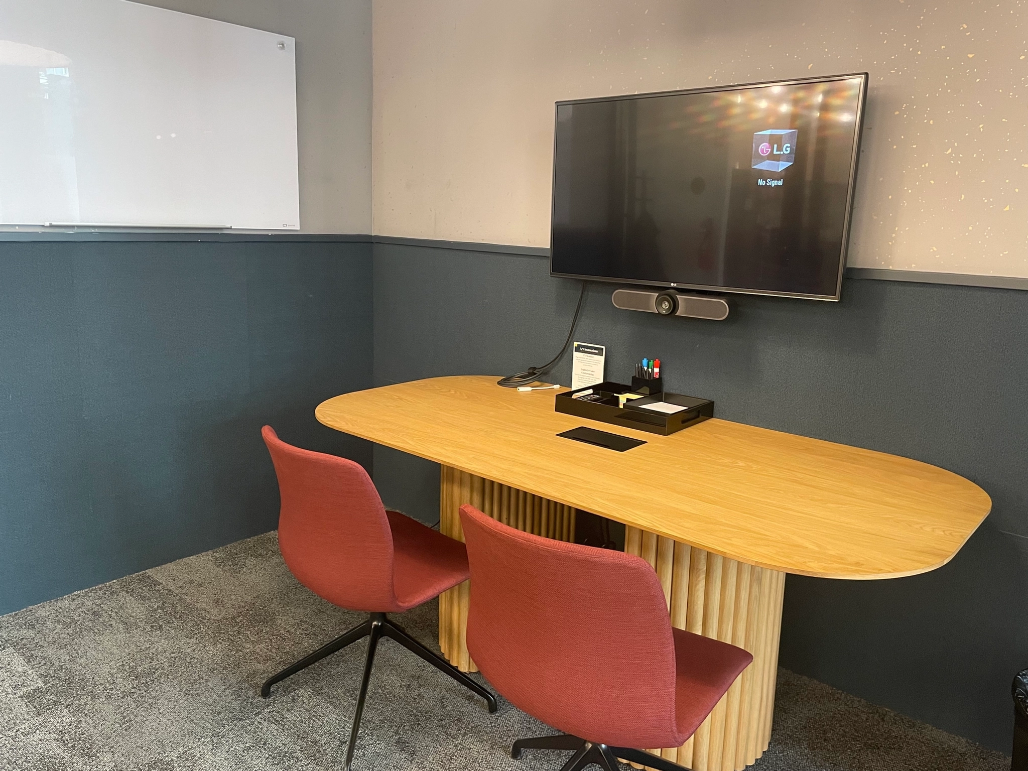 A compact meeting room features a workspace with a wooden table, two red chairs, a wall-mounted TV screen, and a whiteboard.