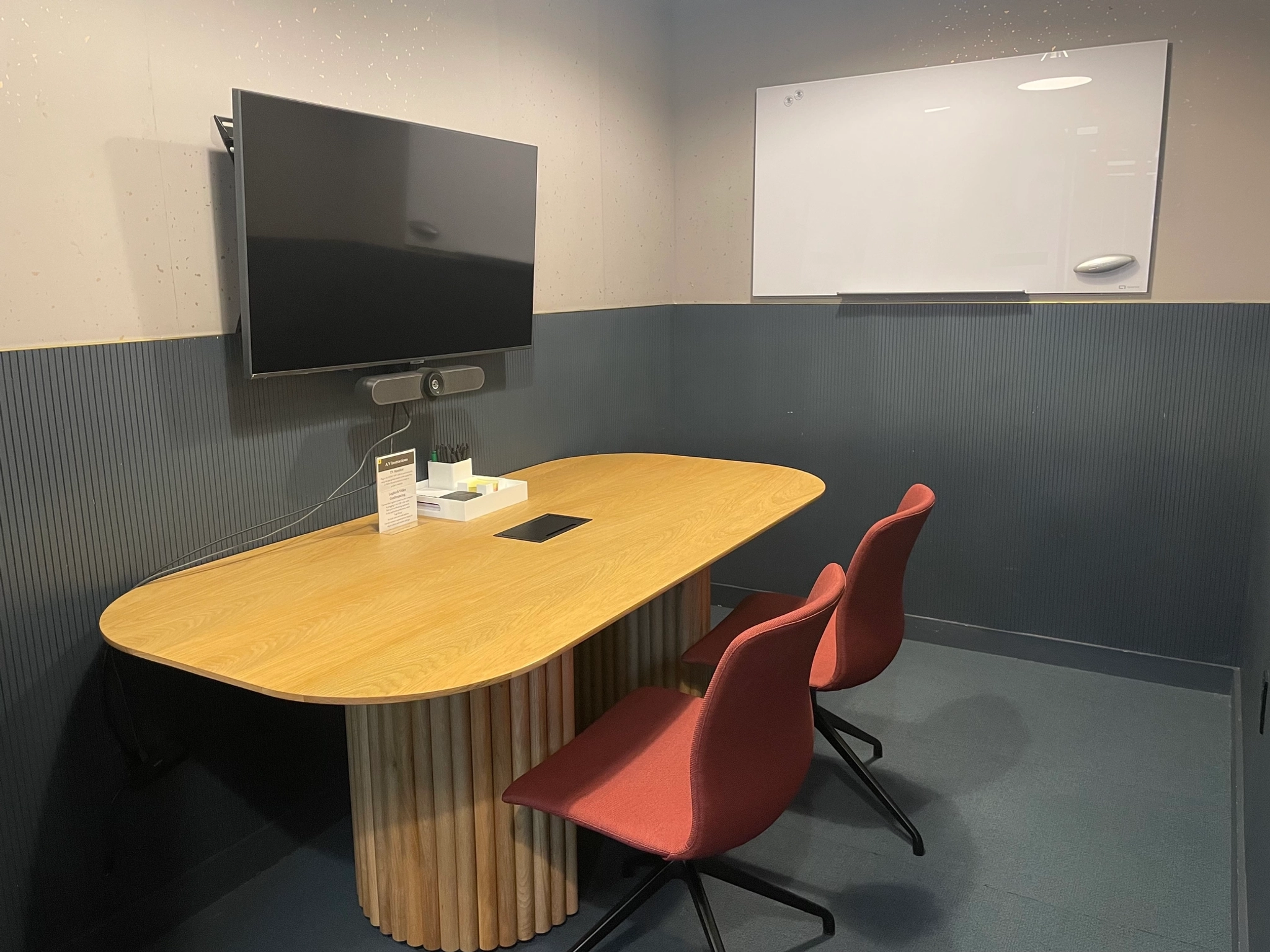 Een compacte kantoorvergaderruimte, compleet met een houten tafel, twee rode stoelen, een aan de muur gemonteerde televisie, een videoconferentiesysteem en een whiteboard, biedt een efficiënte werkruimte.