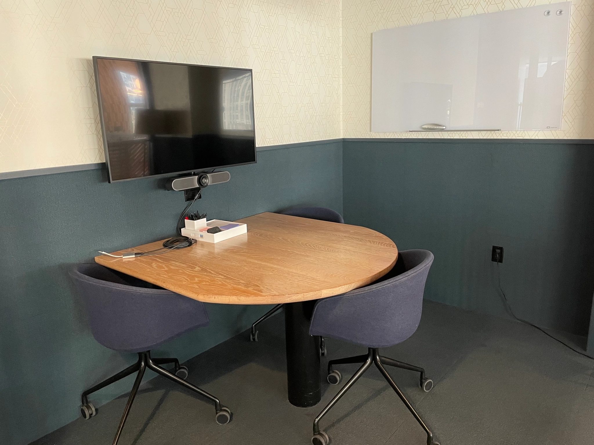 Small coworking meeting room with a round table, two blue chairs, a wall-mounted TV, webcam, and a blank whiteboard—perfect for collaborative workspaces.