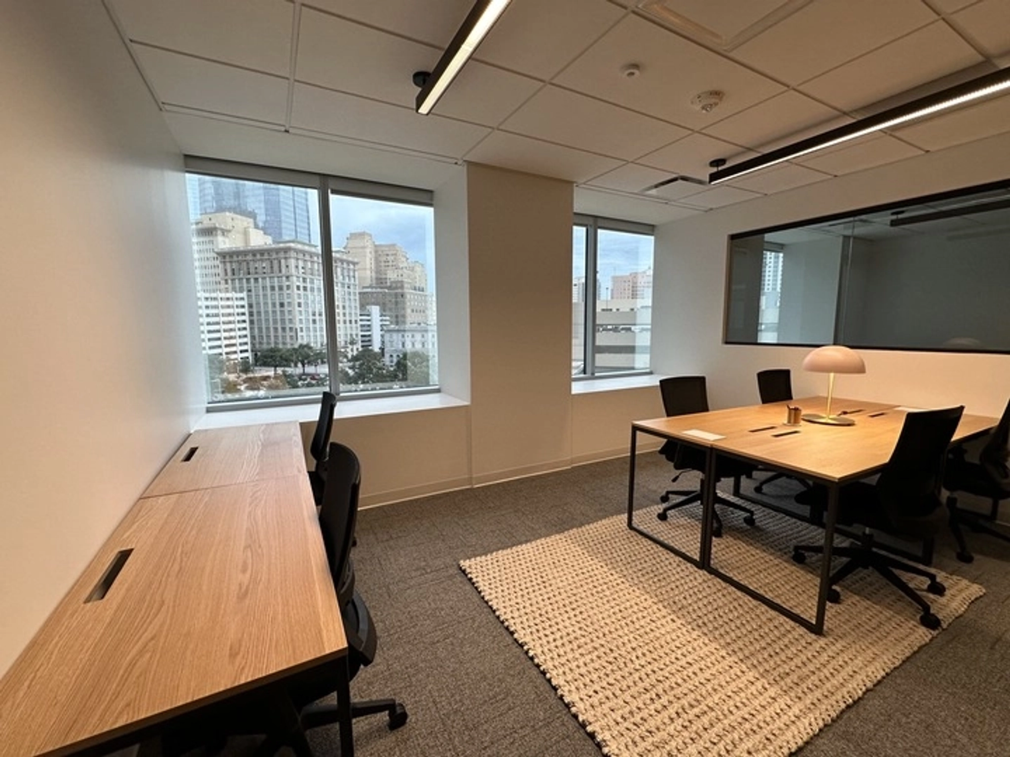 A modern, empty workspace with large windows boasts two wooden desks with chairs positioned along one wall and a table with four chairs on a rug in the center, perfect for a coworking environment. Enjoy the stunning views of city buildings outside.