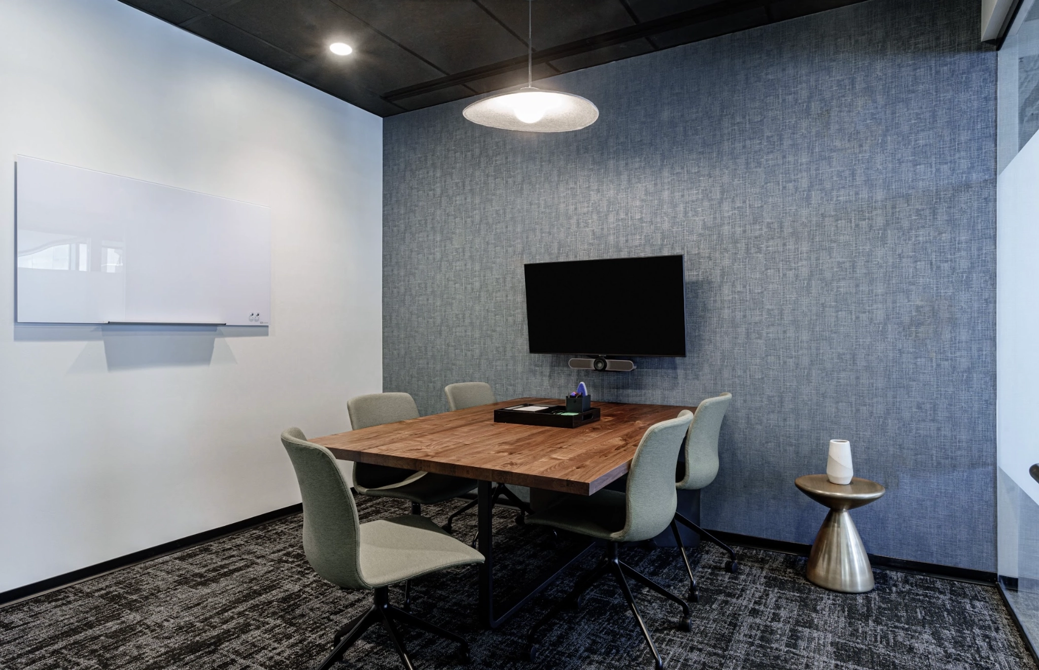 Une salle de conférence minimaliste dans un espace de coworking d'Austin comprend une table en bois, six chaises, une télévision murale, une petite table d'appoint et un tableau blanc.