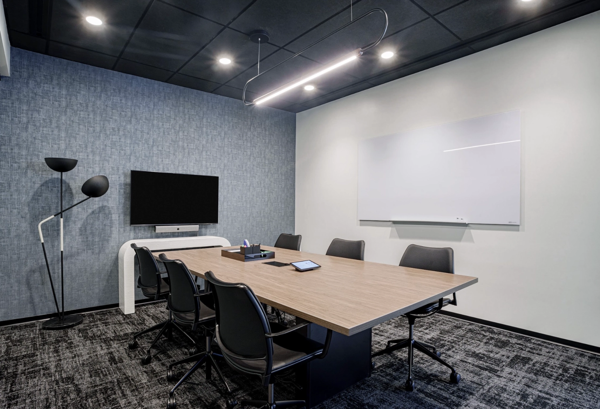 Modern meeting room with a wooden table, six black chairs, a wall-mounted TV, and a whiteboard. The workspace features a black ceiling and blue-gray wall to accent the space.