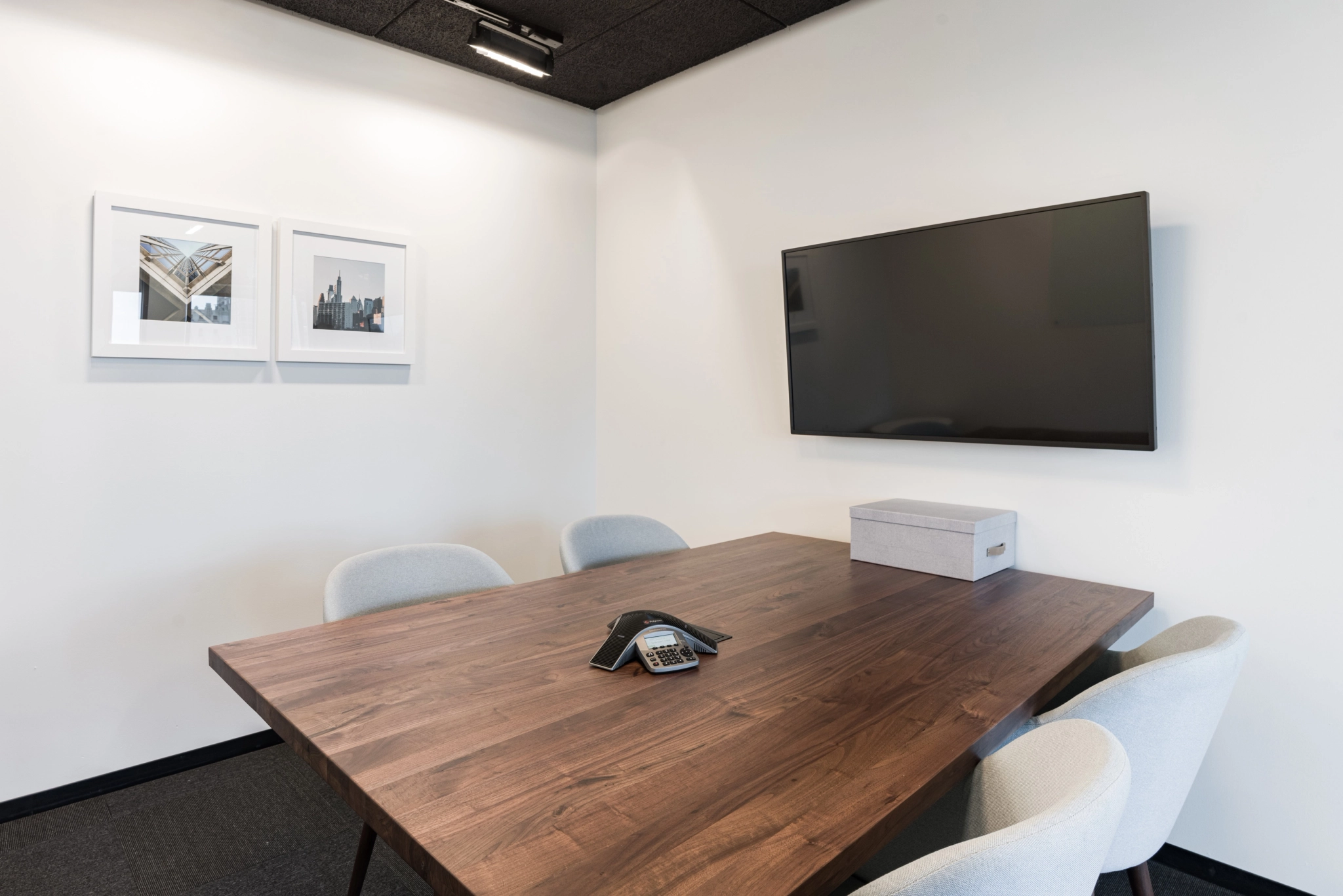Small conference room with a wooden table, four chairs, a phone, and a TV on the wall. Framed pictures reminiscent of Malibu decorate the adjacent wall. A box on the table completes this efficient workspace.