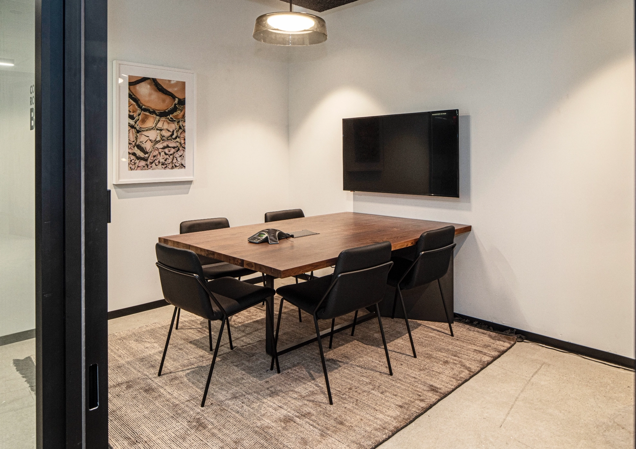 A small meeting room with a wood table, six black chairs, a wall-mounted TV, a light fixture, a phone on the table, and a framed picture reminiscent of Malibu on the wall.