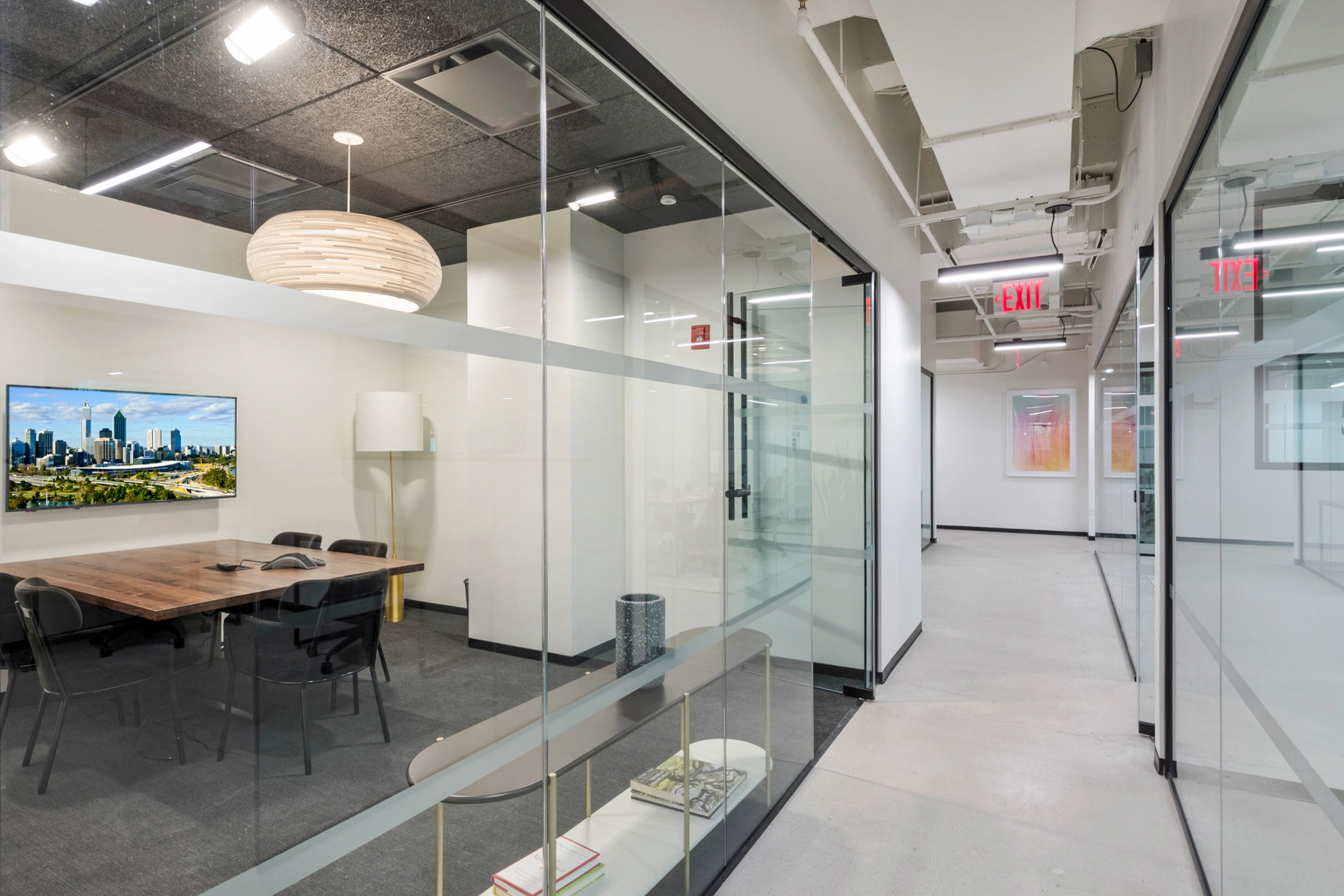 A modern office hallway in a Malibu coworking space features glass-walled conference rooms, a wooden table with chairs, a large wall-mounted TV, and ceiling lights. The exit signs and accent light fixtures are prominently visible.