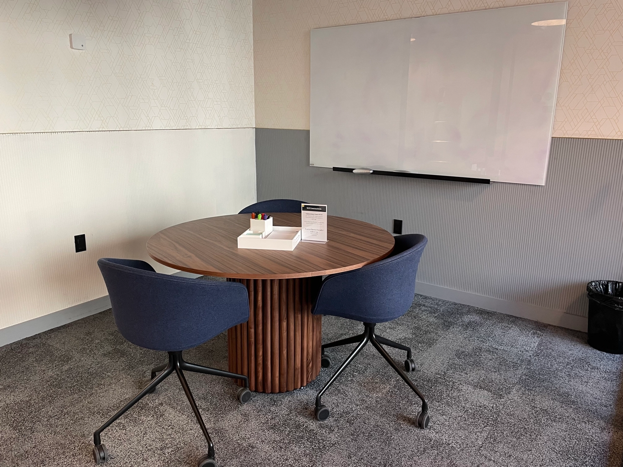 A small office meeting room in New York features a round wooden table, four blue chairs, and a whiteboard on the wall. At the center of this efficient workspace is a small box with pens, ready for brainstorming sessions.