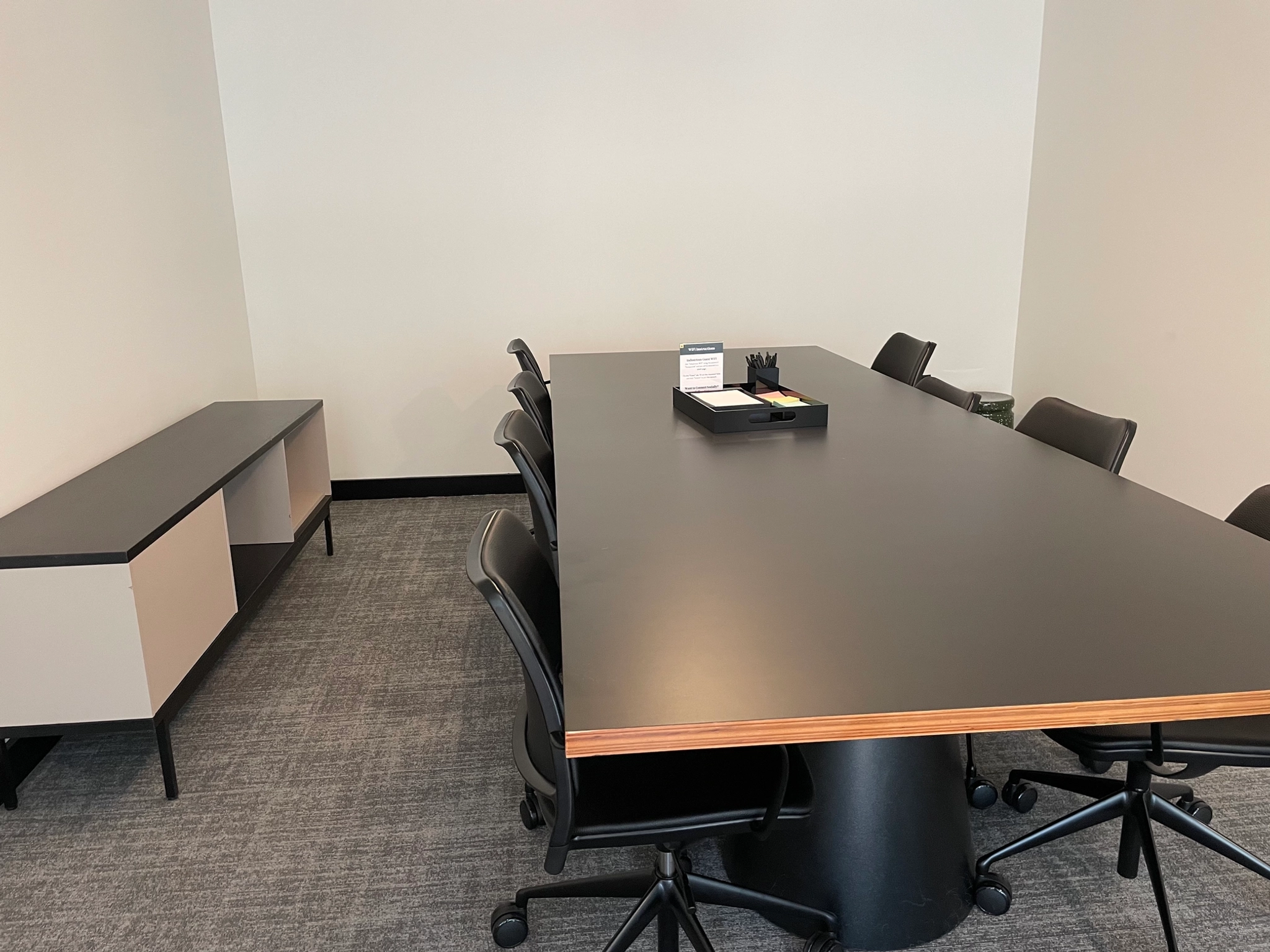 Een moderne werkruimte beschikt over een vergaderruimte met een lange zwarte tafel omringd door acht elegante zwarte stoelen. De inrichting wordt afgerond door een beige zijkast en een dienblad met belangrijke kantoorbenodigdheden.