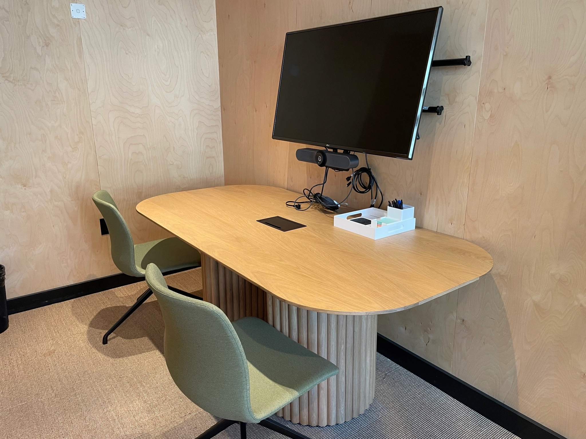 A small office meeting room features a wooden table with two green chairs and a wall-mounted TV screen. The table is equipped with a webcam, cable, and a white tray holding pens, making it an ideal space for coworking sessions.