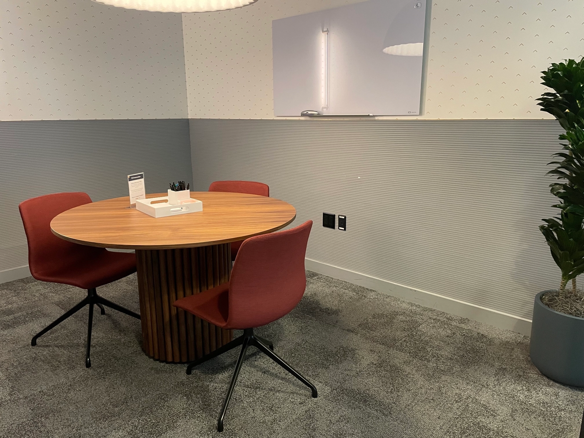 A New York office meeting room featuring a round wooden table, four red chairs, a whiteboard on the wall, and a potted plant in the corner.