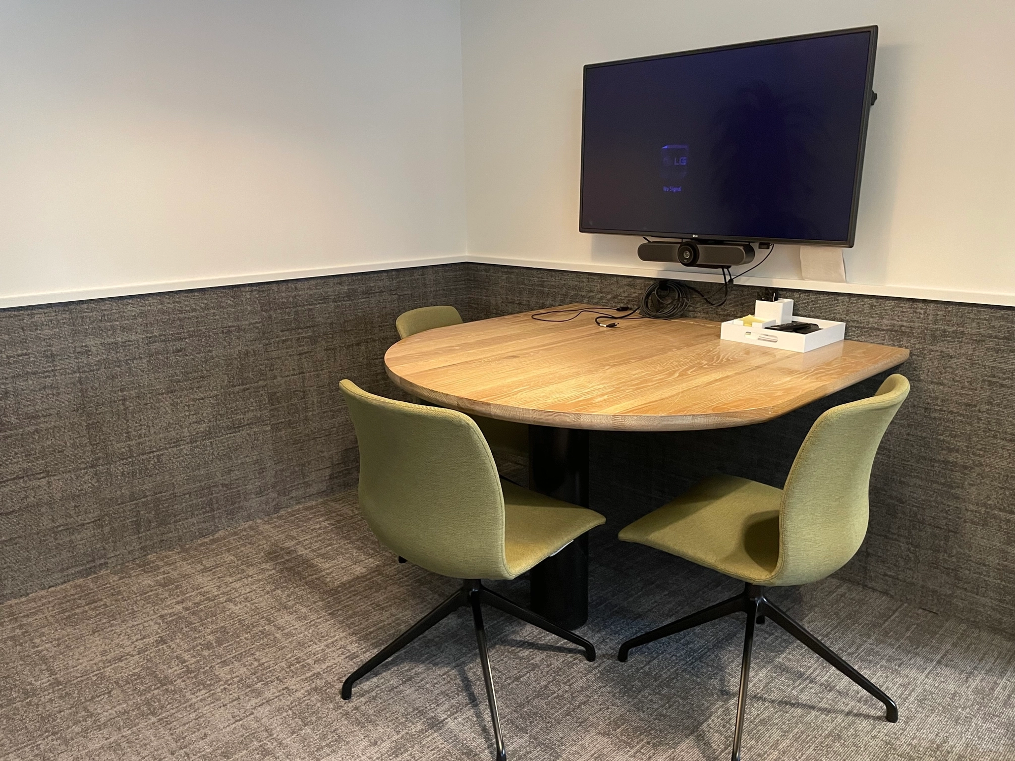 A cozy office meeting room with a round wooden table, three green chairs, and a wall-mounted TV screen equipped with a video conferencing camera, perfect for coworking sessions.