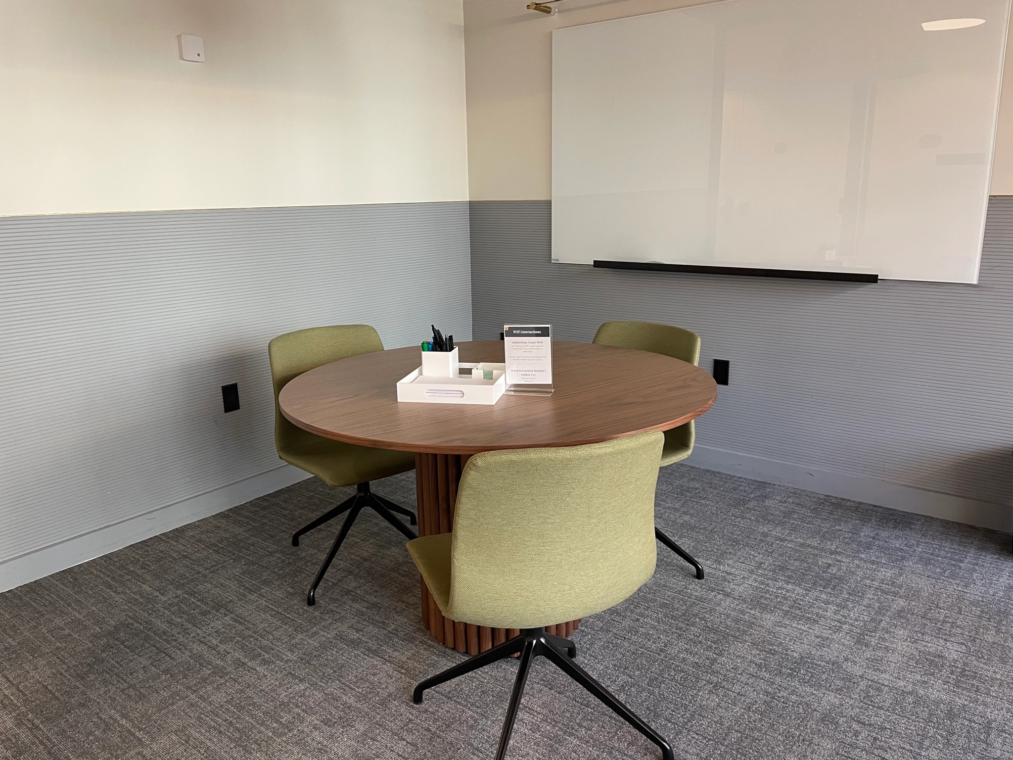 A small office meeting room in New York features a round wooden table, three green chairs, a whiteboard on the wall, and office supplies neatly arranged on the workspace.