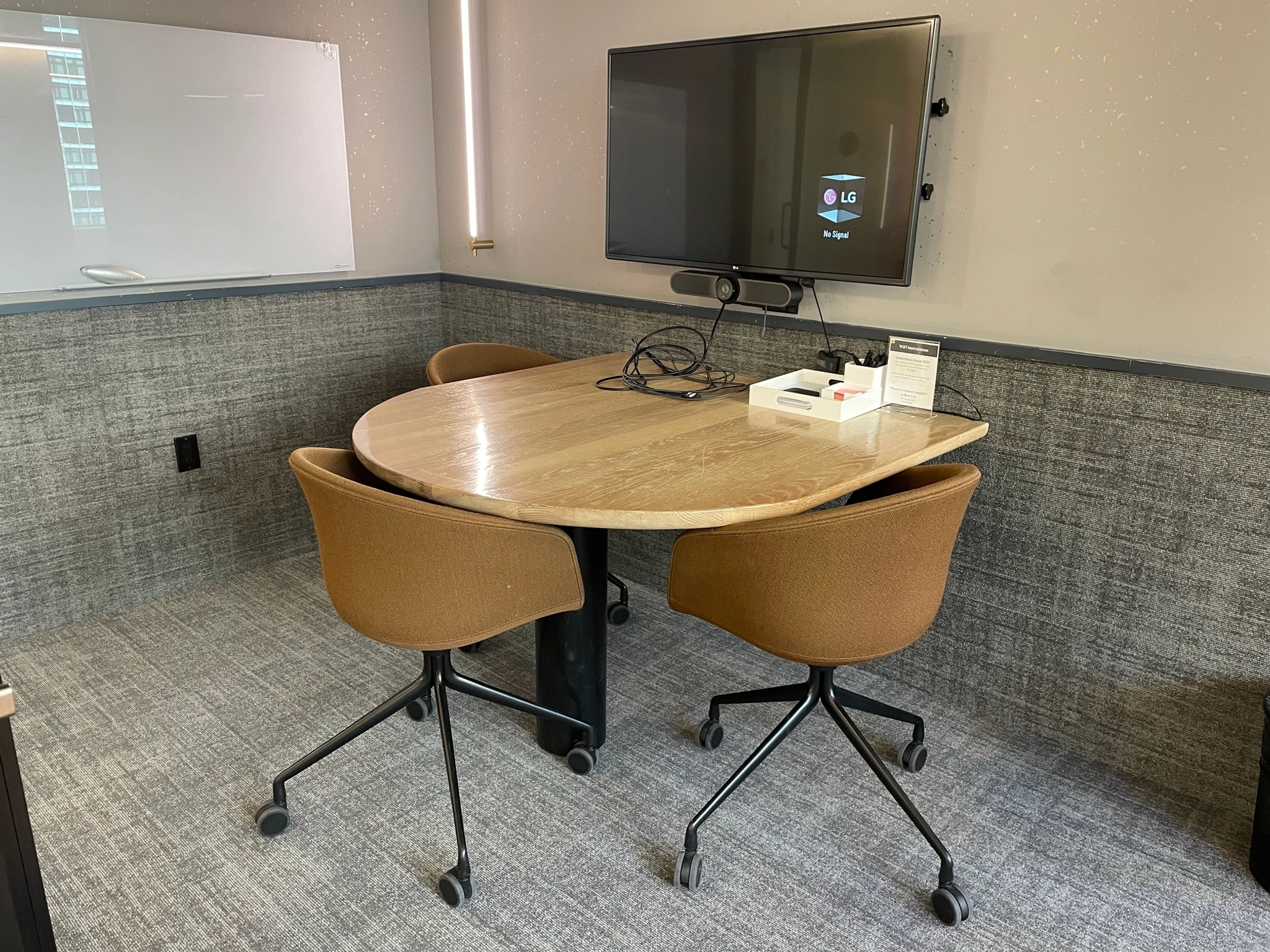 A cozy workspace featuring a round wooden table, three brown chairs on wheels, a TV on the wall, and a whiteboard in the corner. This adaptable office setting is perfect for brainstorming sessions or collaborative coworking discussions.