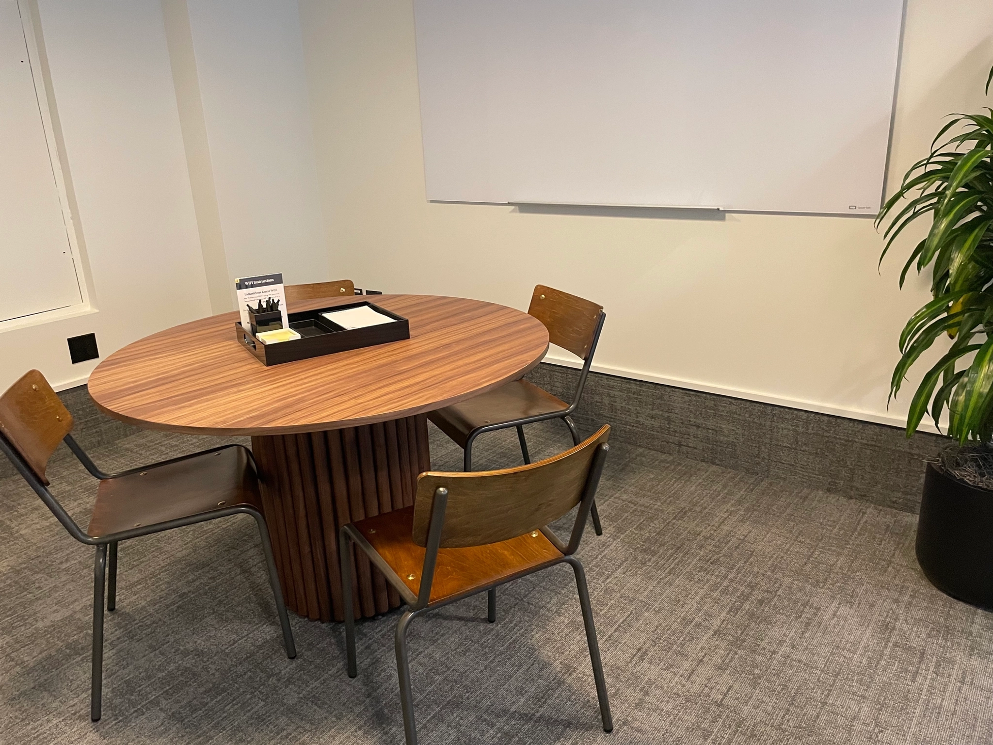 A small coworking space featuring a round wooden table, four chairs, a whiteboard on the wall, and a potted plant in the corner.