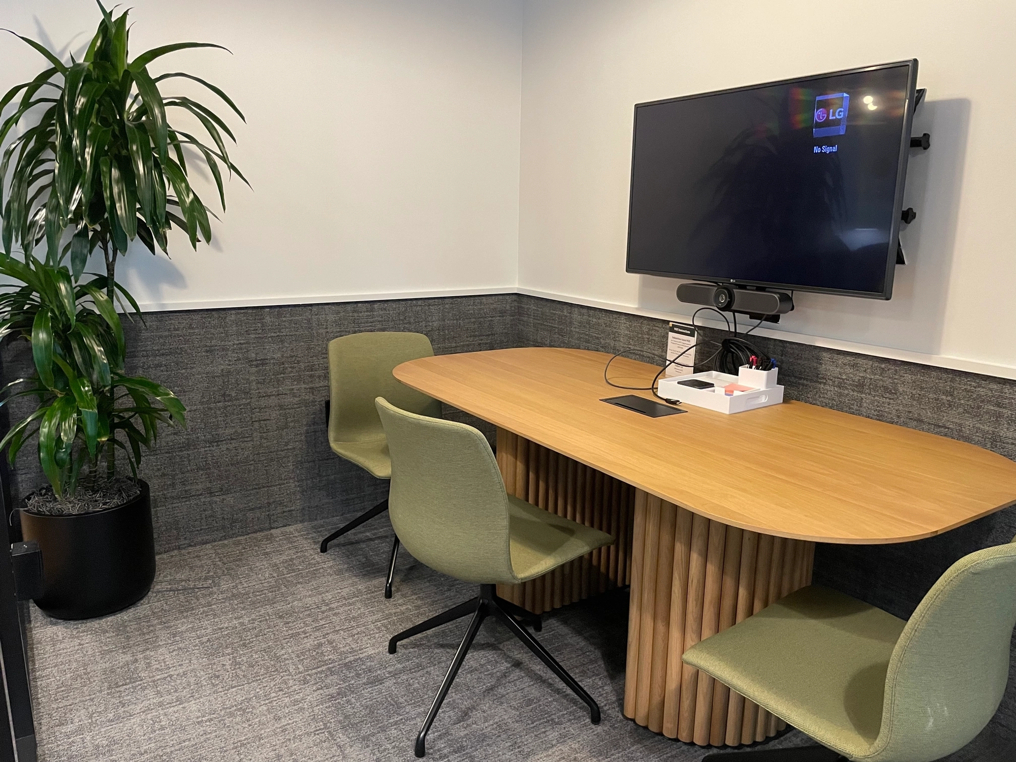 This cozy office meeting room features a wooden table, three green chairs, a wall-mounted monitor, and a potted plant in the corner, creating an inviting workspace.