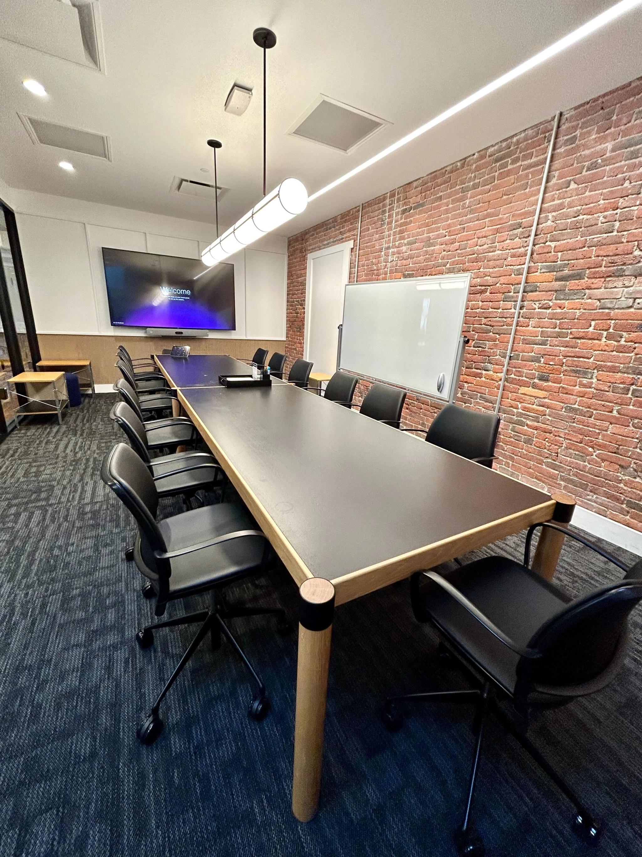 A modern meeting room with a long table, ten chairs, a wall-mounted TV, a whiteboard, and an exposed brick wall offers the perfect workspace for collaboration.