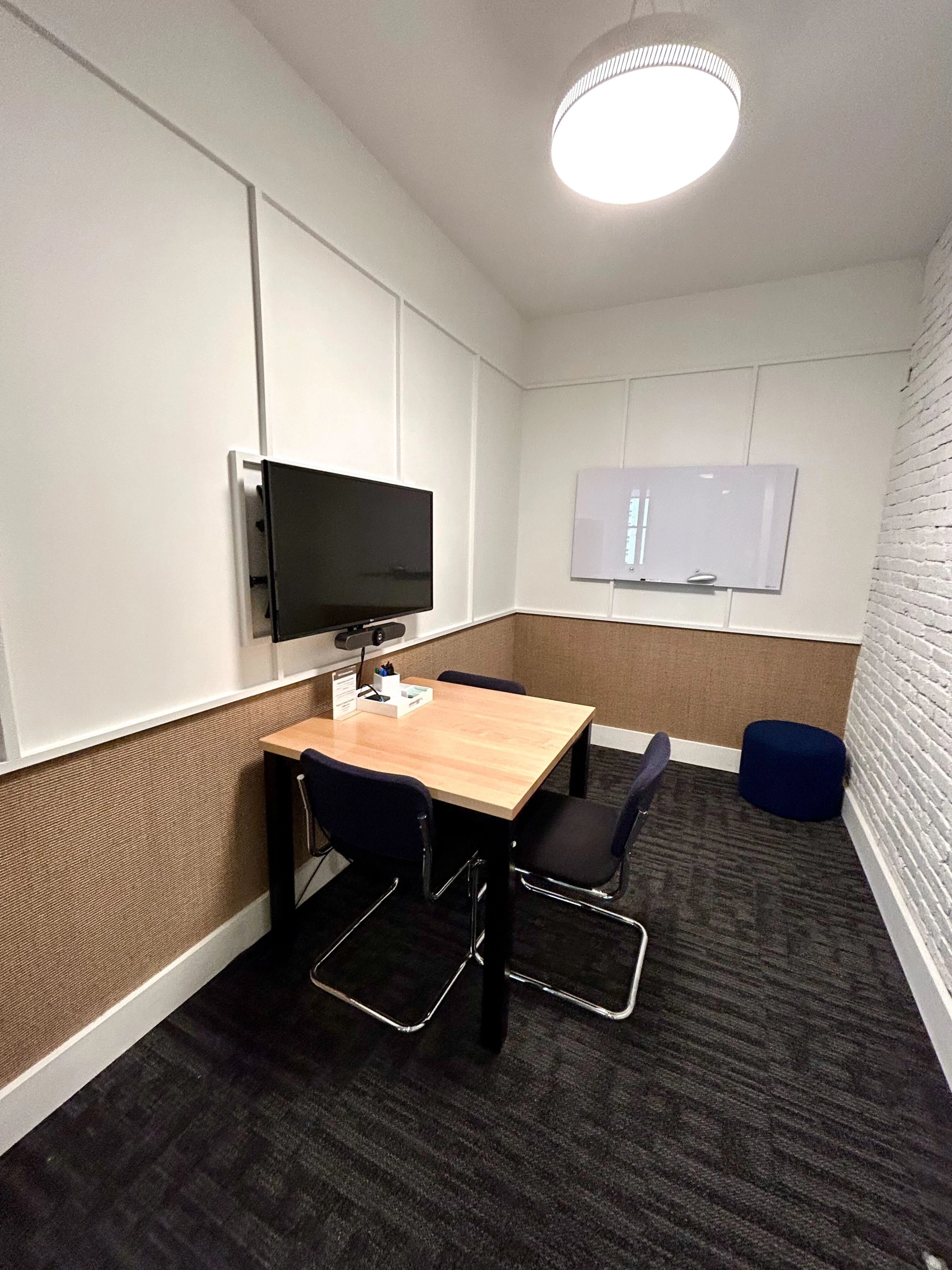 A small coworking meeting room in Boston features a table, two chairs, a wall-mounted TV, a whiteboard, and a black pouf. The room is cozy with its carpeted floor and white brick wall.