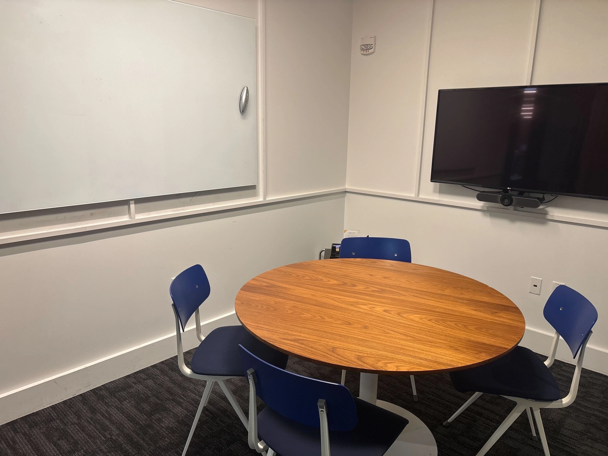 A small Boston meeting room with a round wooden table, four blue chairs, a wall-mounted whiteboard, and a flat screen TV—perfect for productive office gatherings.