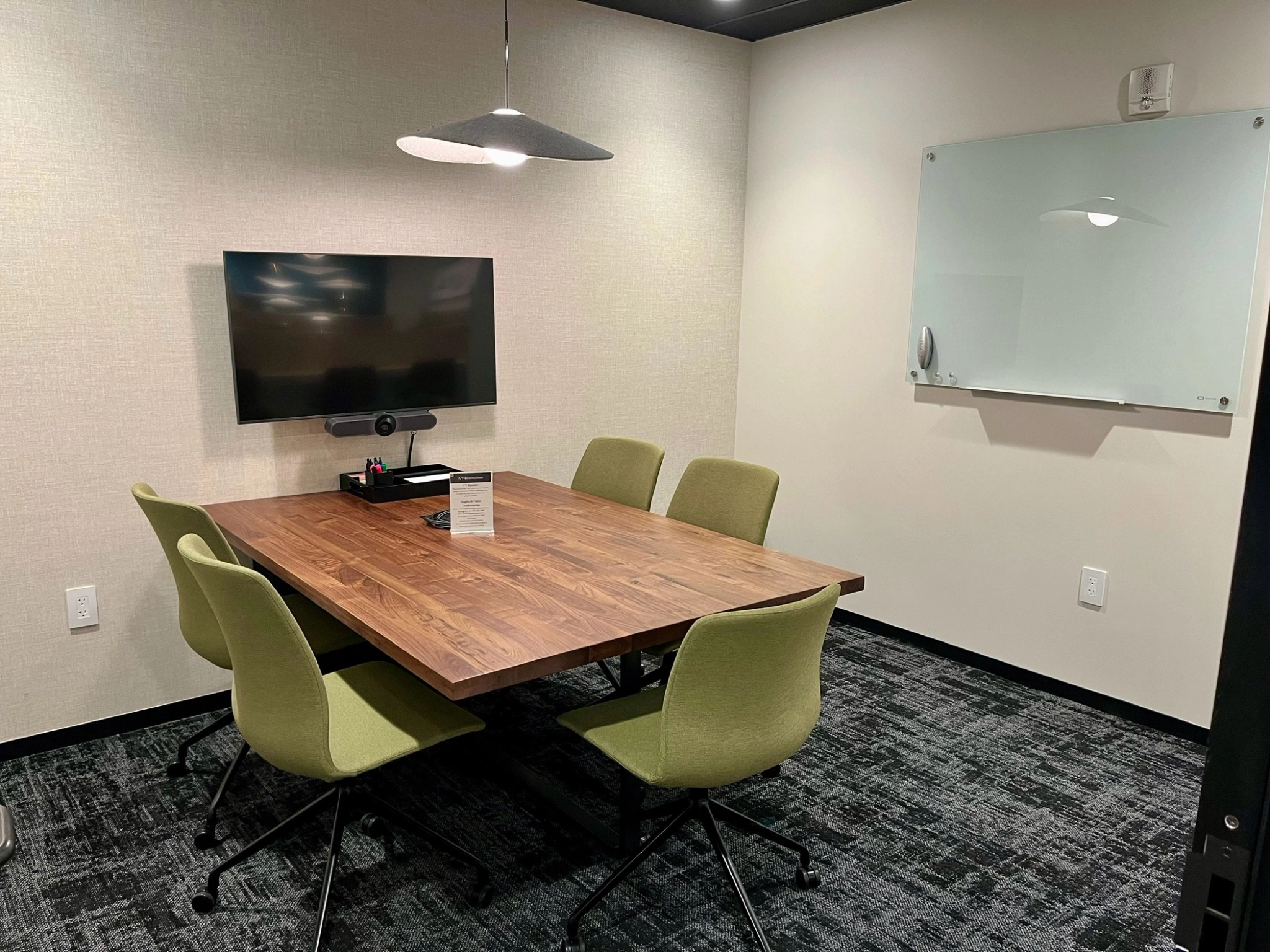 A small conference room in a coworking space with a wooden table, four green chairs, a wall-mounted TV, a video conferencing device, and a whiteboard.