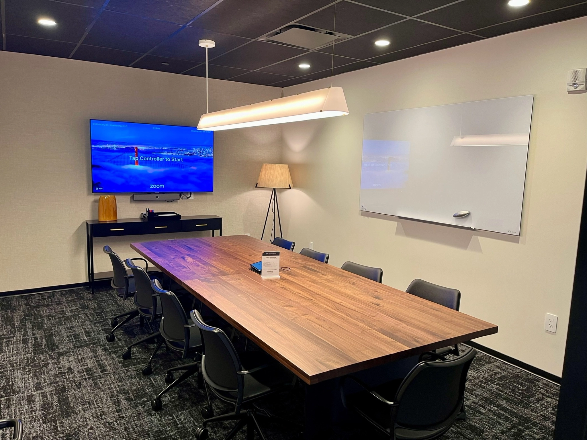 A conference room in Coconut Grove featuring a wooden table, eight chairs, a large screen displaying a Zoom welcome screen, a whiteboard, and a standing lamp provides an ideal workspace for collaboration.