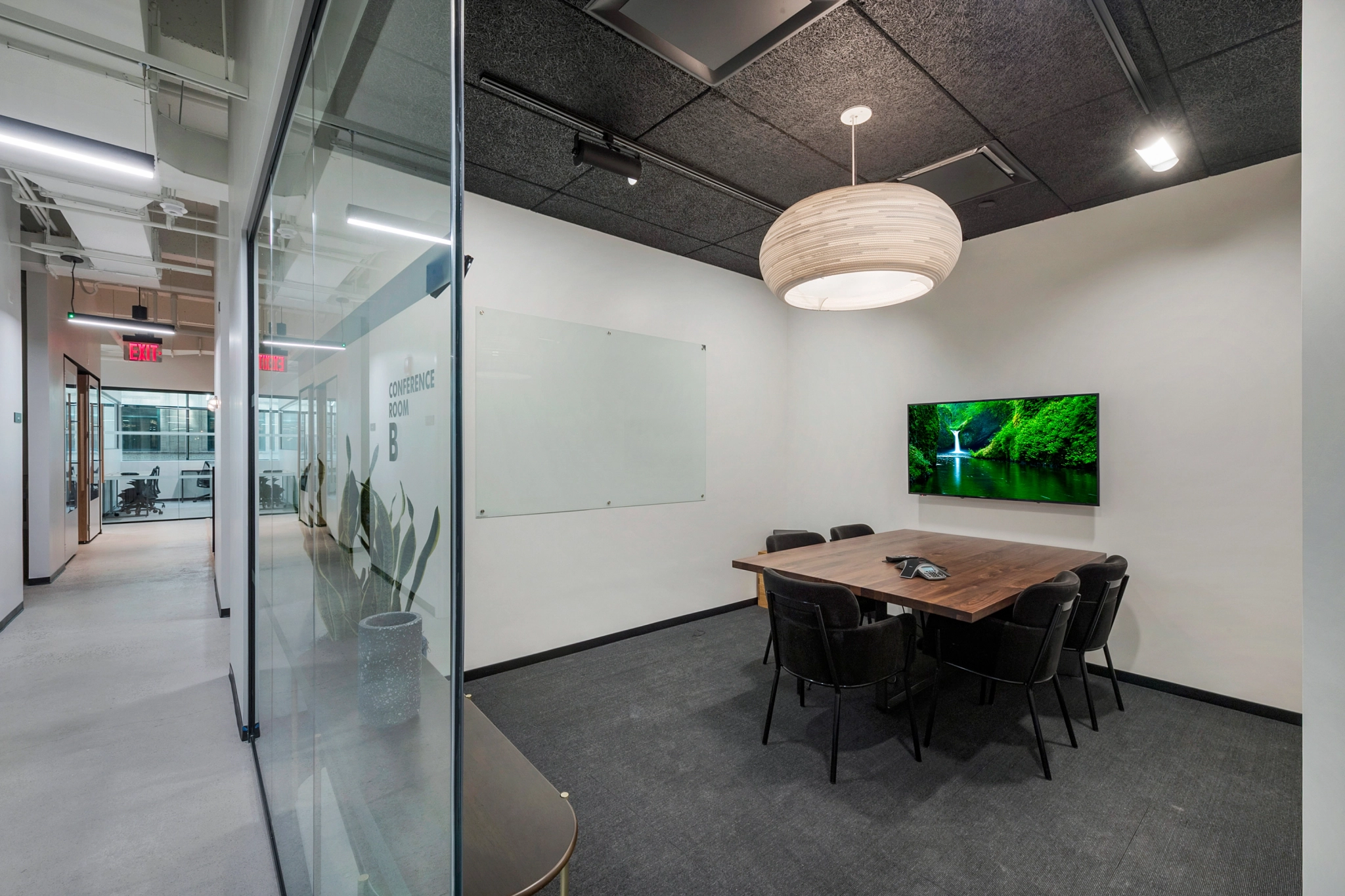 A small, modern meeting room with a wooden table, black chairs, a wall-mounted TV displaying a nature scene, a whiteboard, and a round ceiling light. Glass walls separate it from the hallway, ideal for coworking sessions in the office.