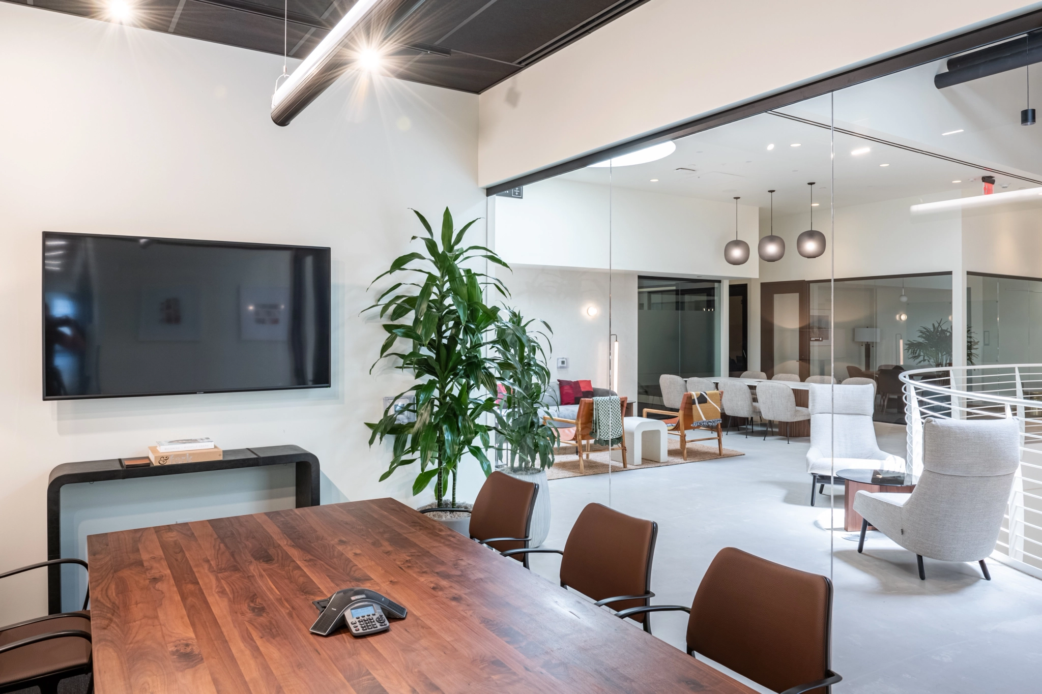 A modern meeting room in Los Angeles featuring a wooden table, brown chairs, a wall-mounted TV, and a potted plant. A glass wall separates this stylish workspace from the lounge seating in the background.