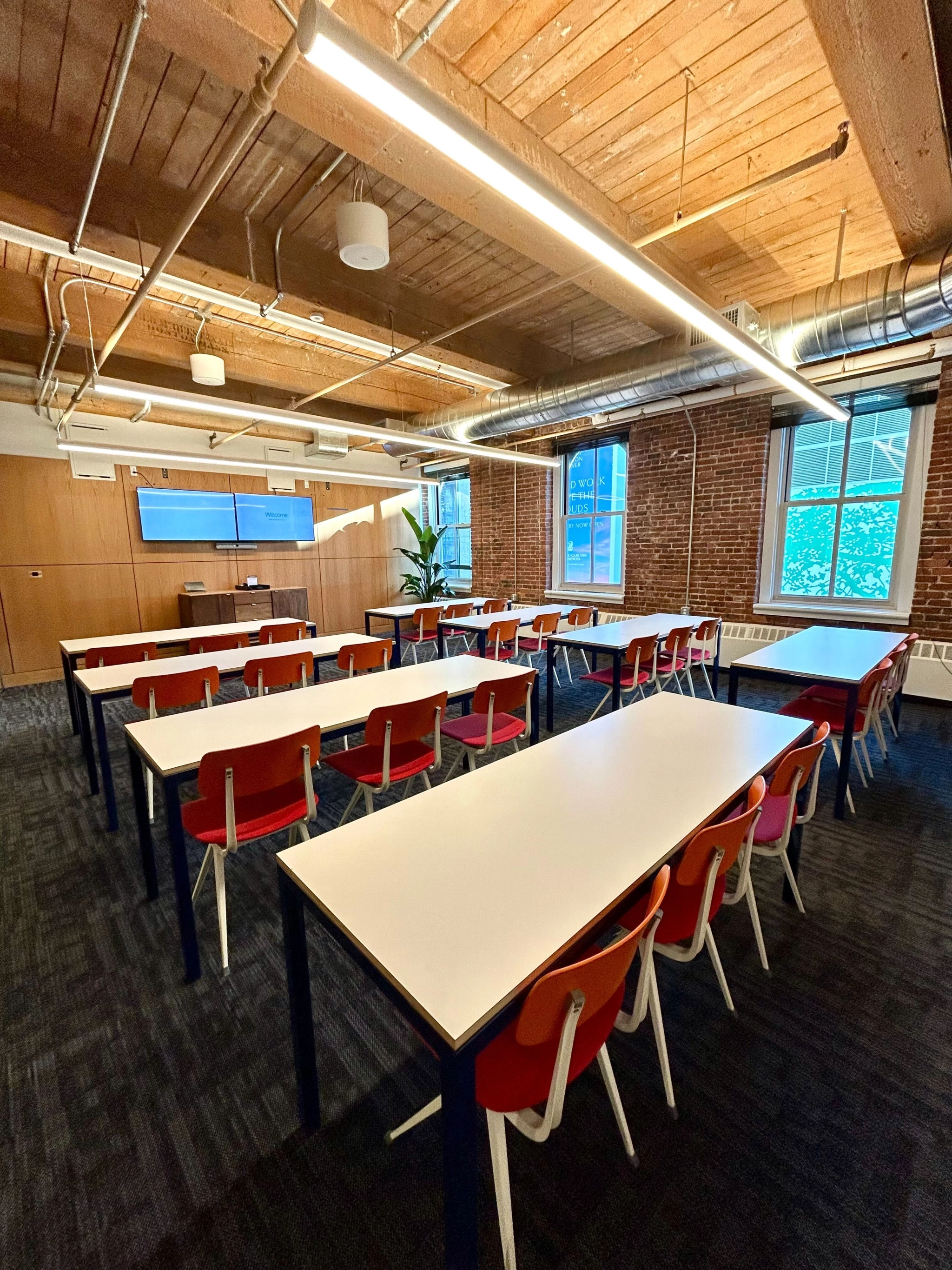 A modern workspace featuring rows of white tables and red chairs, complemented by a large window, wooden ceiling, and industrial-style lighting. Perfectly blending the essence of an inspiring coworking environment with functionality.