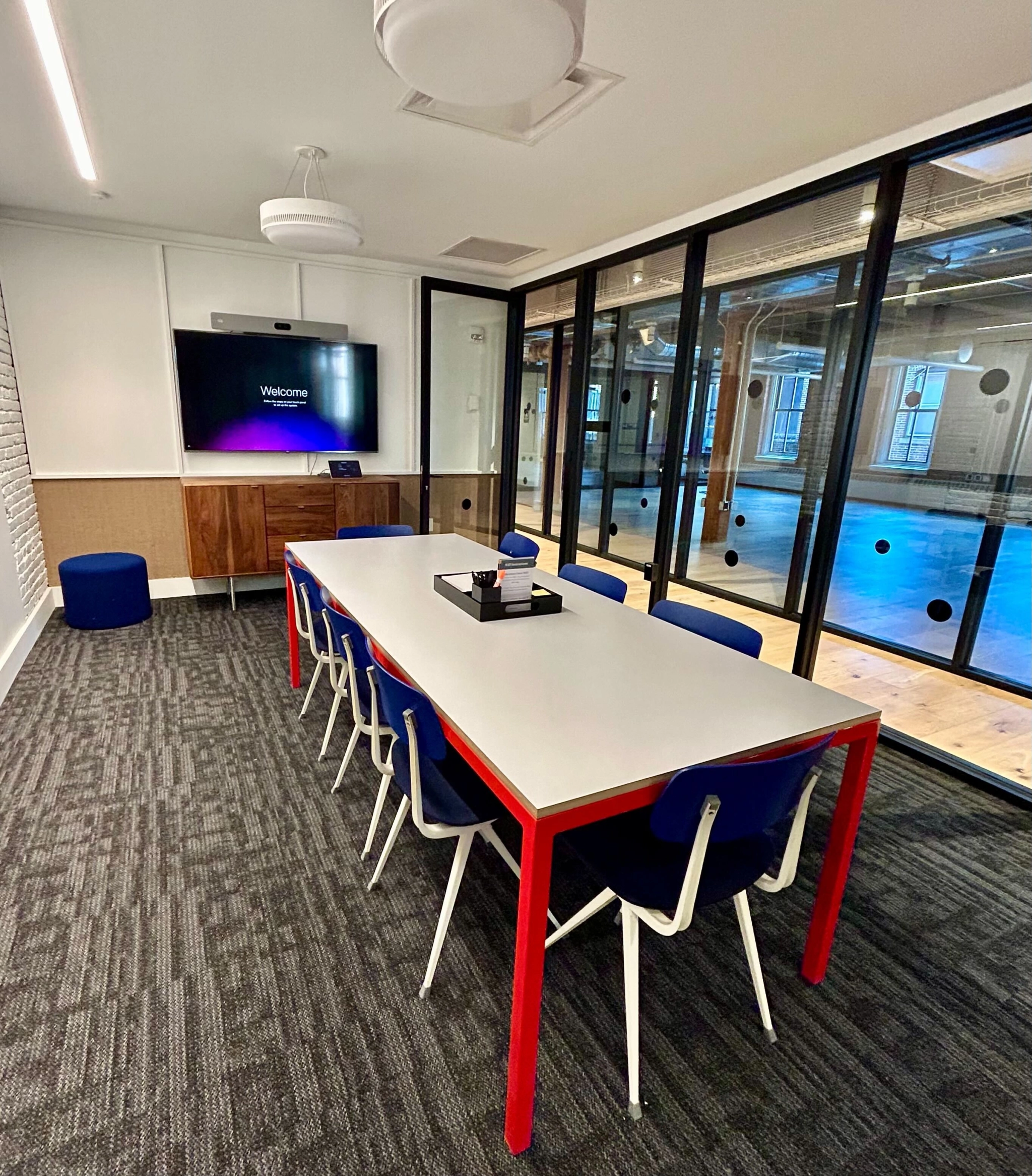 The office meeting room features a long white table, blue chairs, a wall-mounted screen, and large glass windows overlooking an open workspace.