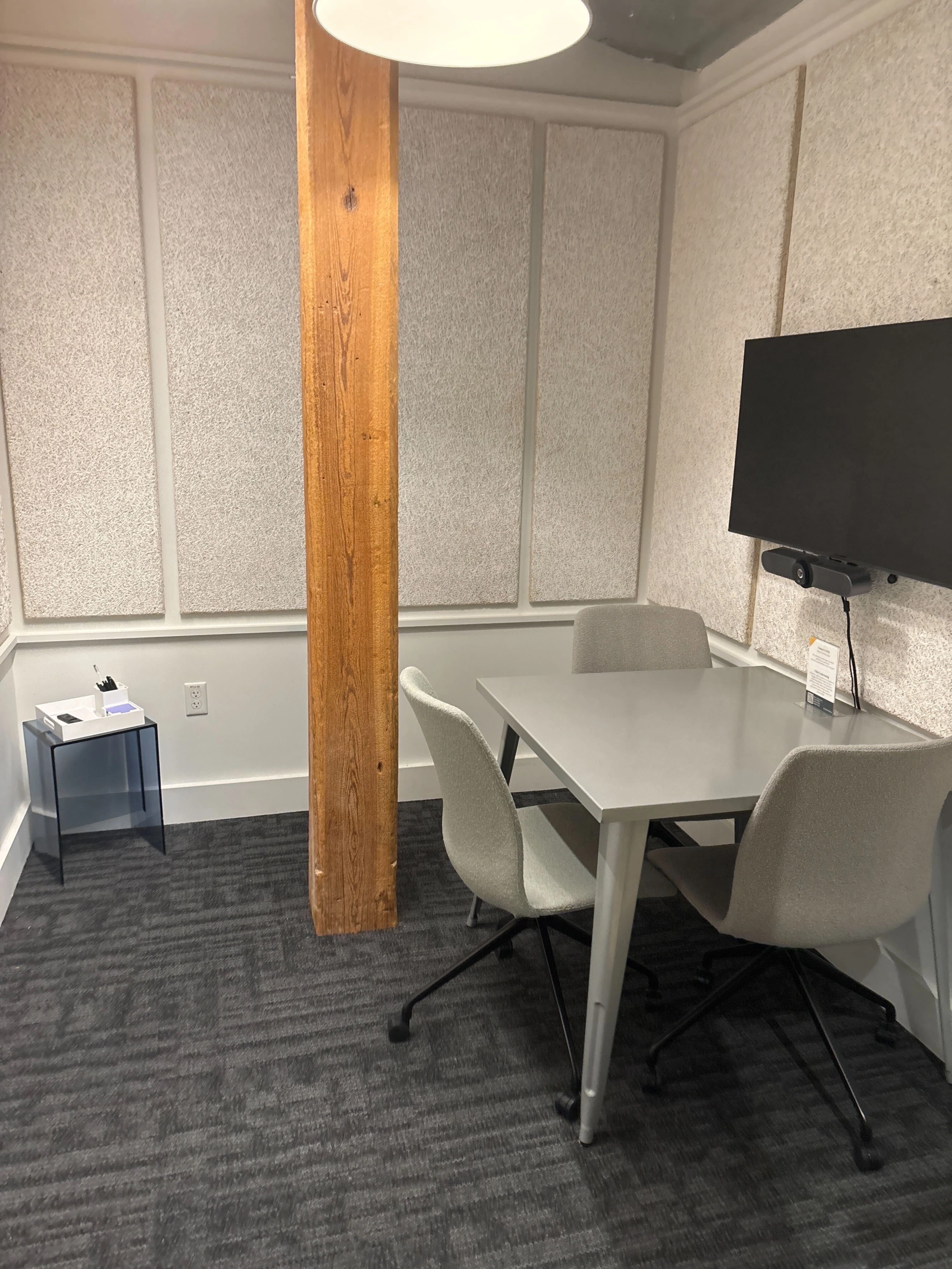 A small coworking office with a table, three chairs, a wall-mounted monitor, a wooden pillar, and a side table stocked with tissues and sanitizer.
