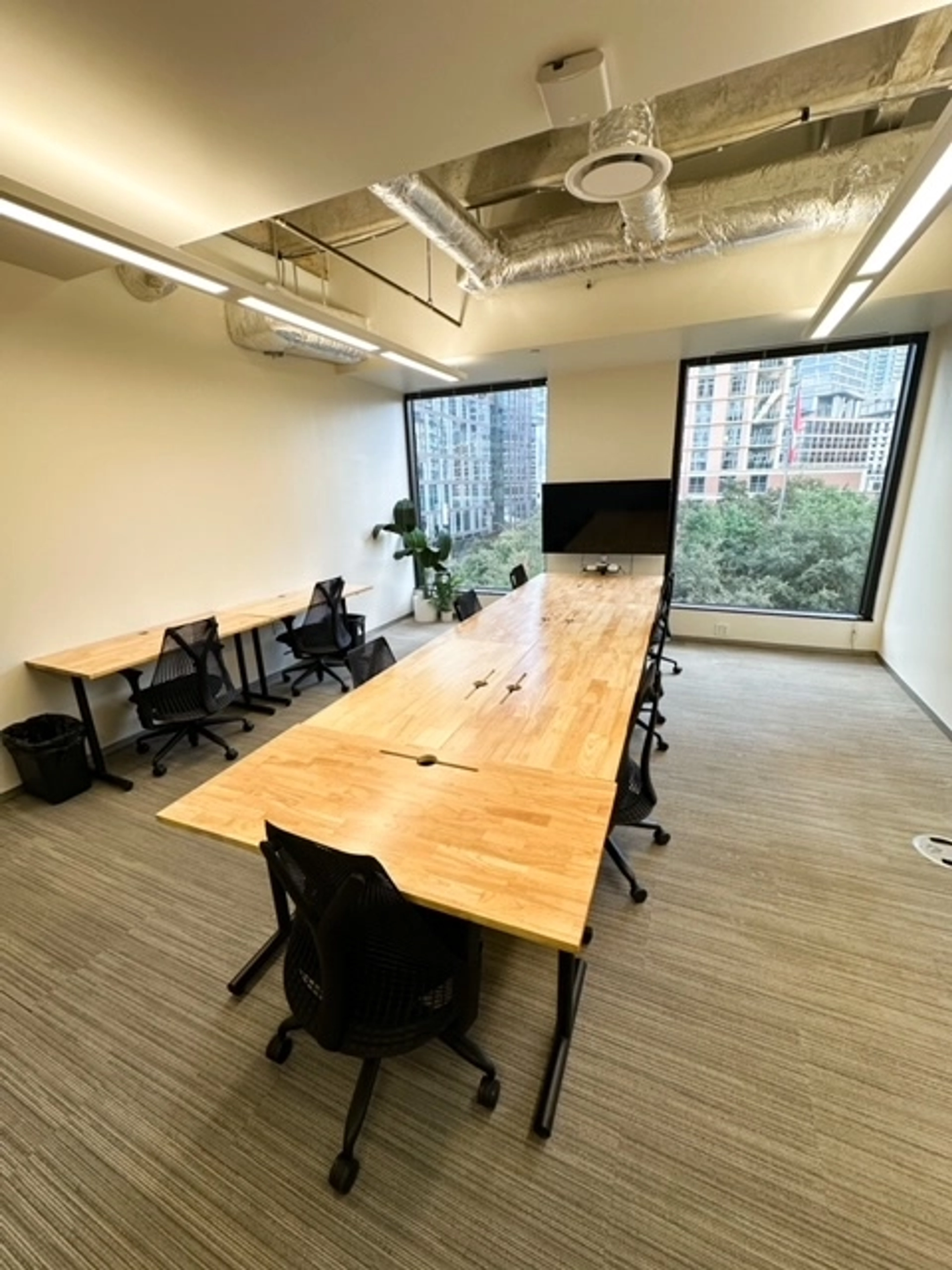 Ein Büro in Austin mit einem langen hölzernen Konferenztisch und mehreren Stühlen ist ein idealer Besprechungsraum. Mit zwei Schreibtischen an der Wand, großen Fenstern für Tageslicht und einem Fernsehbildschirm auf der Rückseite ist es perfekt für gemeinsame Coworking-Sitzungen.