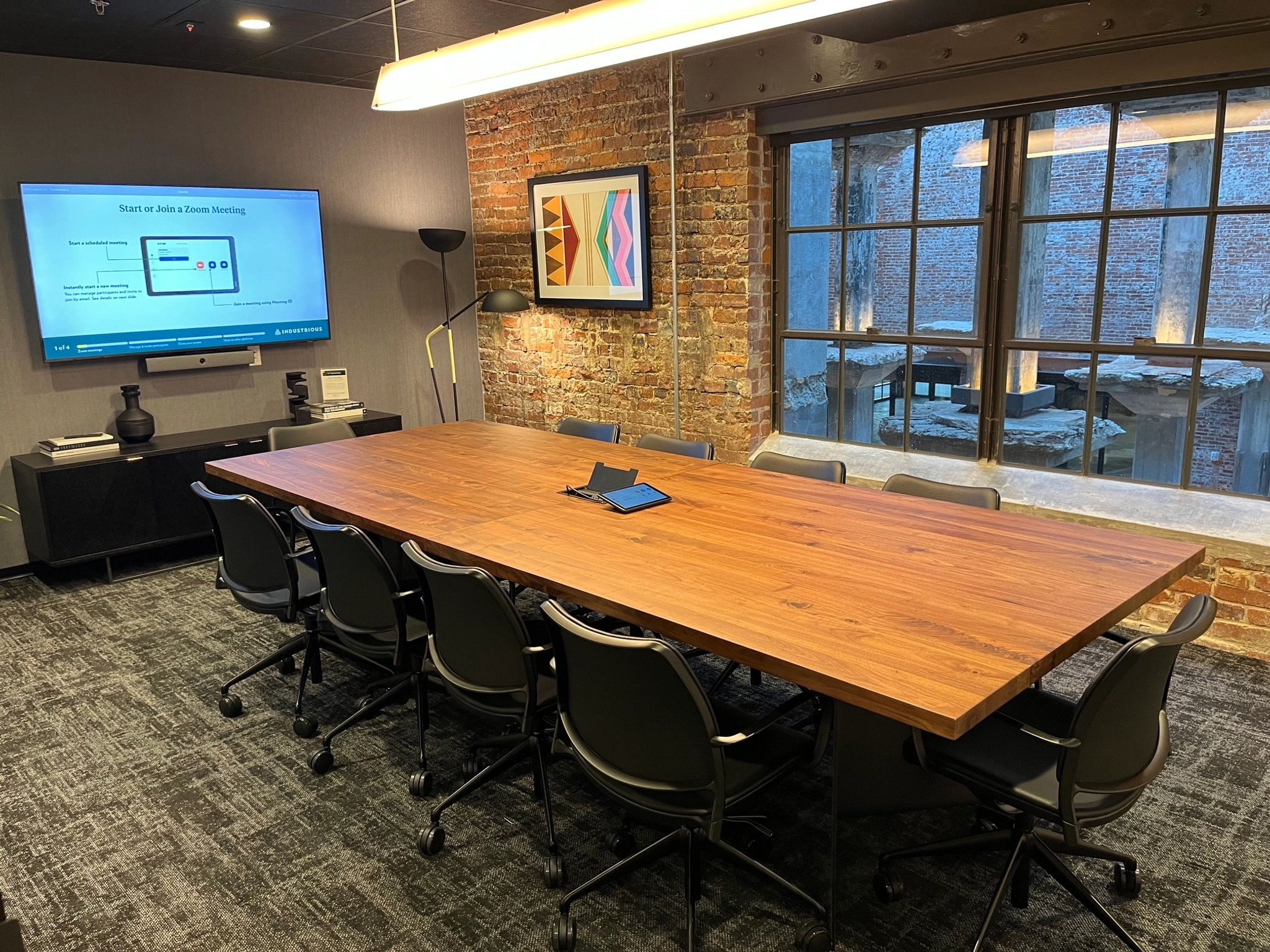 A sophisticated meeting room with a long table, nine chairs, and a large monitor displaying a meeting screen. The exposed brick walls add character to this dynamic workspace.