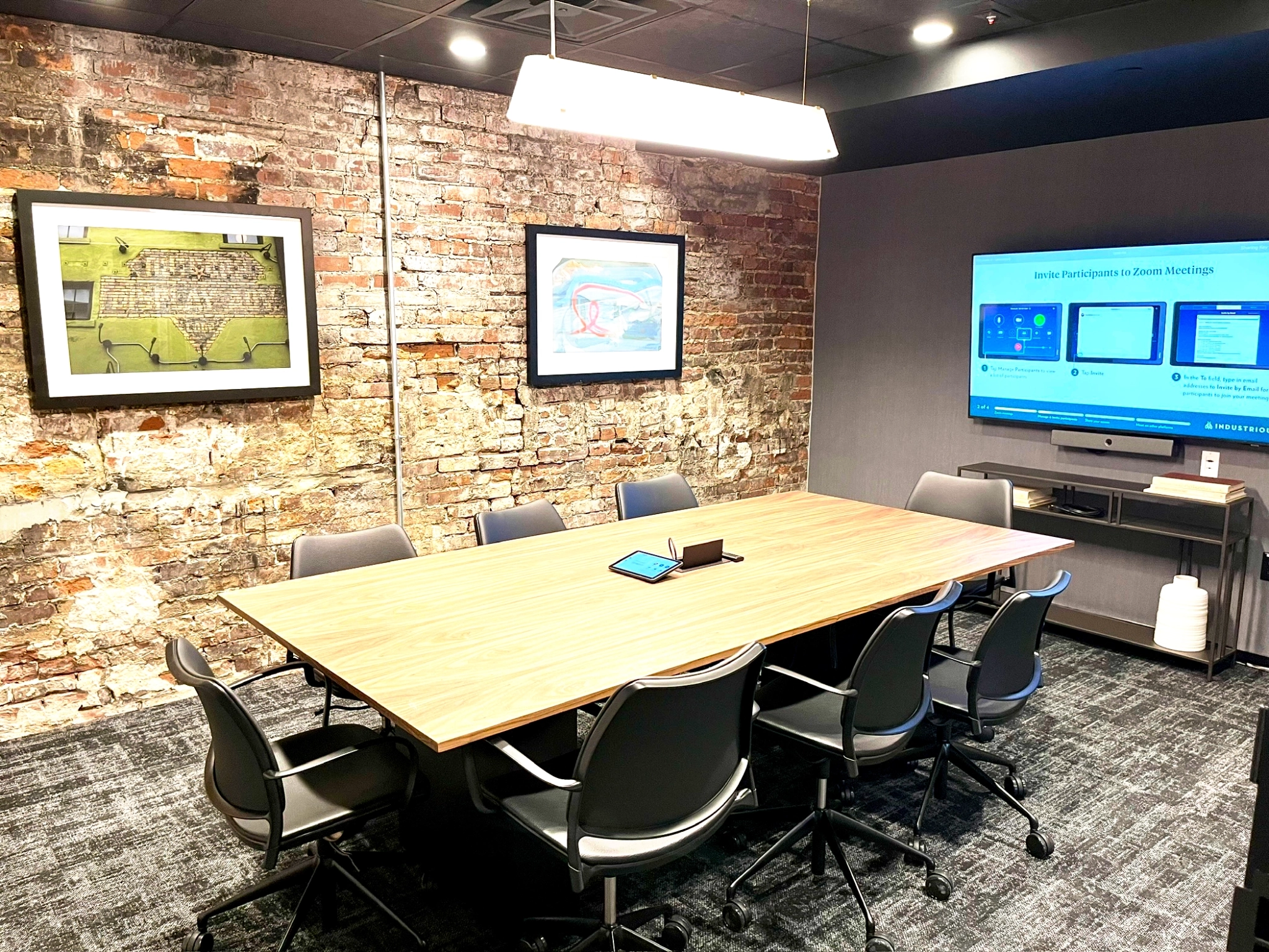 The Nashville conference room features a wooden table, eight chairs, an exposed brick wall adorned with framed artwork, and a TV screen displaying a Zoom interface. A tablet sits on the table, adding to the sleek functionality of this stylish meeting room.