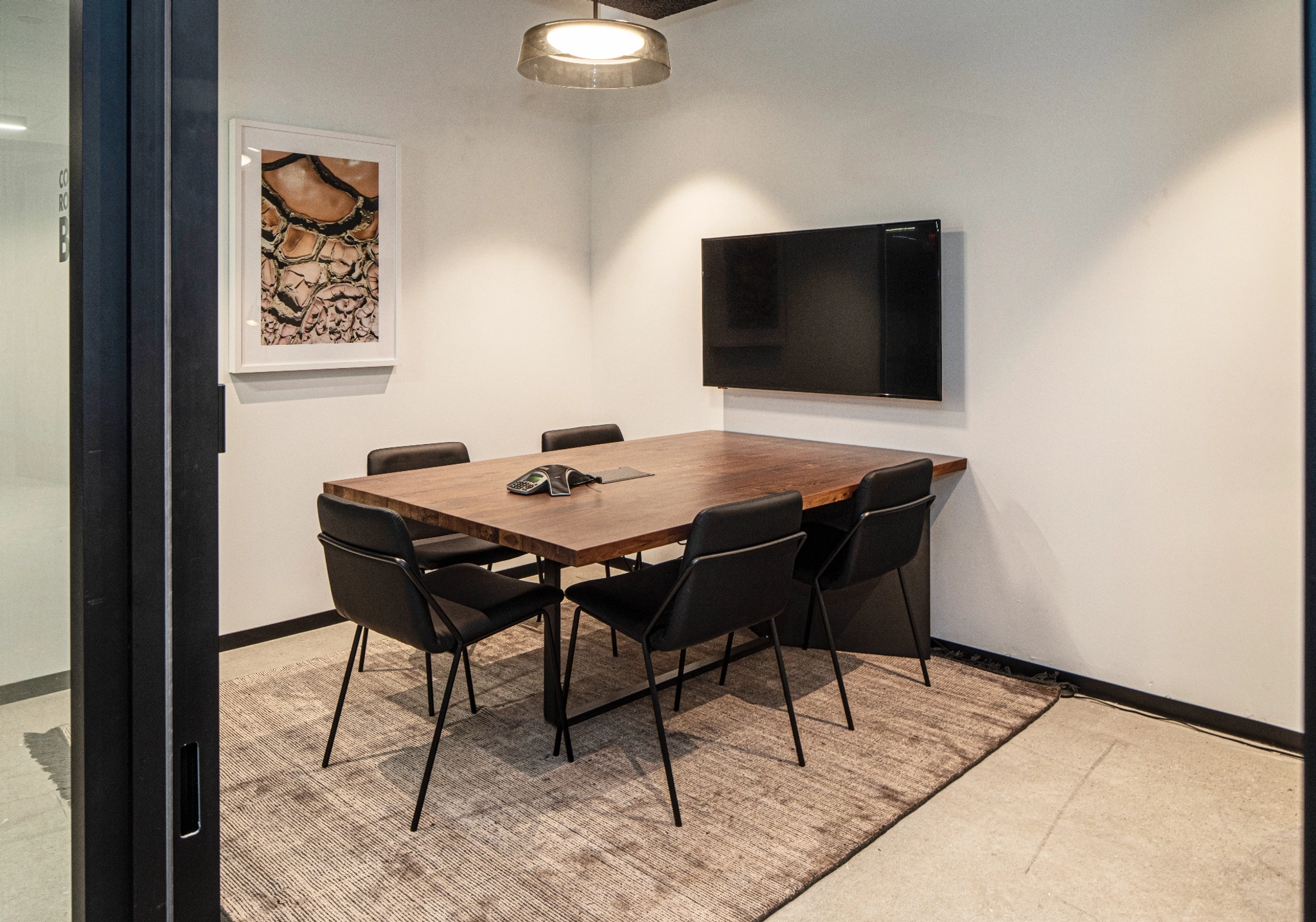 A coworking space featuring a wooden table, six black chairs, and a wall-mounted TV. This meeting room has white walls adorned with framed artwork, complemented by a stylish rug and ceiling light.