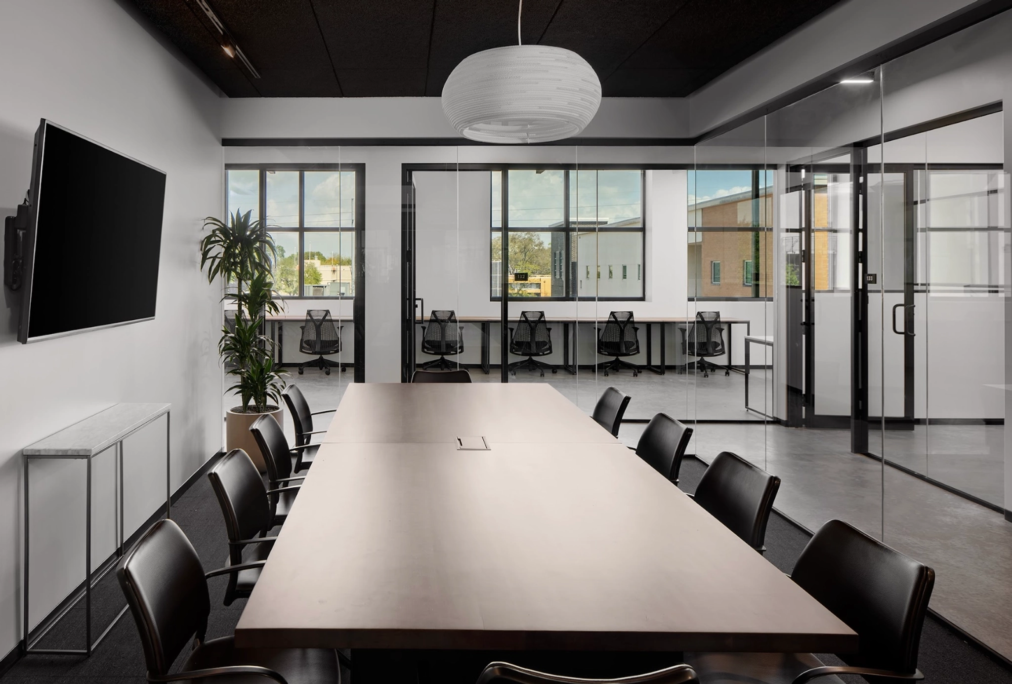 A modern meeting room featuring a long table, black chairs, and a wall-mounted TV is illuminated by large windows. The sleek design flows seamlessly into the workspace beyond, where office cubicles with chairs are visible through elegant glass partitions.