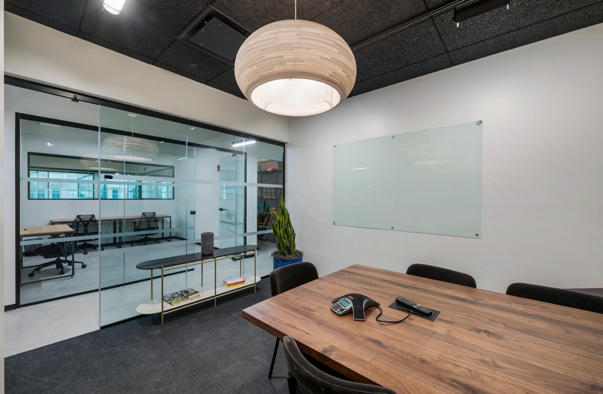 A sleek Santa Monica meeting room featuring a wooden table, chairs, phone, and whiteboard under a stylish hanging lamp. It opens to a glass-walled workspace filled with desks and chairs.