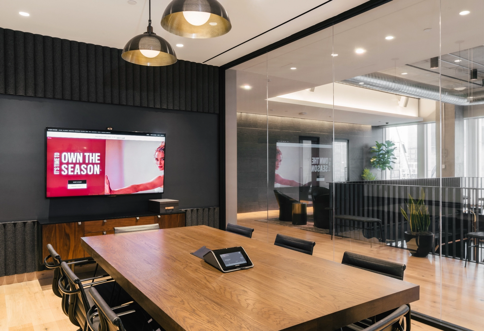 A modern workspace featuring a wooden table, black chairs, and a wall-mounted screen displaying "Own the Season." This sleek office boasts large windows filling the meeting room with natural light.